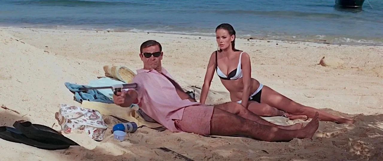 Sean Connery is seen lounging on a sandy beach, wearing a light pink short-sleeved shirt and matching shorts. He is reclining casually, propped up on one elbow, and sporting dark sunglasses. Beside him is a woman in a black and white bikini, sitting upright with a composed expression. The setting is a sunny day by the ocean, with clear blue water visible in the background. The mood is relaxed yet slightly tense due to Sean Connery holding a distinctive prop—a spear gun—adding an element of intrigue and action to the scene. The beach setting is accentuated by items like a colorful beach towel, a floral bag, a bottle of sunscreen, and a pair of flippers lying nearby. The lighting is bright and natural, typical of a sunny day, enhancing the vivid colors of the sand and sea. This image evokes themes of leisure mixed with suspense.