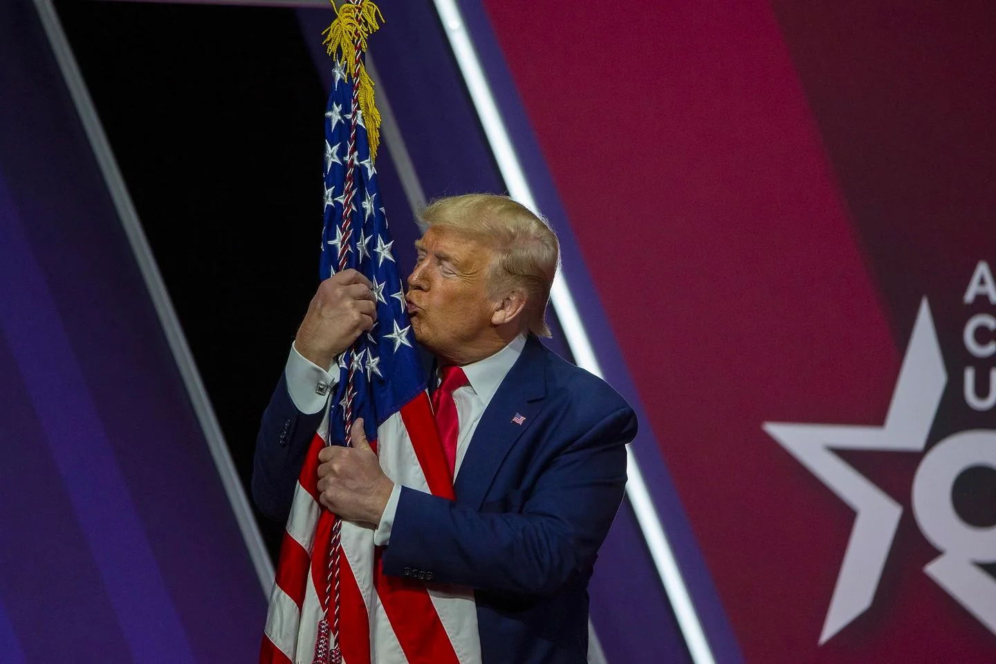 1) Rich description: The image shows Trump at a public event, embracing and kissing an American flag. He is dressed in a dark blue suit with a white shirt and a red tie, projecting a formal and patriotic demeanor. The background features a stylized star design with the partial text "AC" and "USA," likely indicative of the event's theme or sponsor. The setting is indoors, with a stage and a decorative backdrop that uses bold colors such as purple and red, adding to the ceremonial atmosphere. The flag is held tightly by Trump, and his expression is one of affection and reverence. The lighting is bright and focused, illuminating Trump and the flag prominently, creating a focal point for the audience and the photograph. The composition centers on Trump's interaction with the flag, symbolizing a display of national pride.
2) On-screen text block:
- Title: "AC USA"