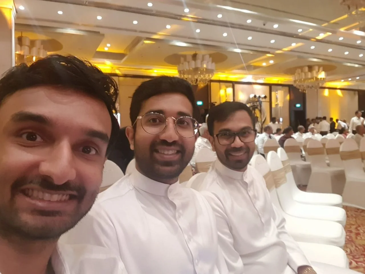 In the image, three individuals are seated together in what appears to be a formal event setting, characterized by an indoor venue with elegant decor. The lighting is warm and inviting, with chandeliers and ceiling lights casting a golden hue. The attendees, including Lehan Edirisinghe and Chadu Sumathipala, are dressed in traditional white attire, suggesting a culturally significant or formal occasion. They are seated on white chairs adorned with beige sashes, indicating a well-organized setup. The individuals are smiling and appear to be enjoying the event, adding a cheerful and relaxed mood to the scene. The background shows other attendees and a setup of cameras, likely capturing the event's proceedings. The atmosphere is lively, with people engaged and attentive to the ongoing activities.
