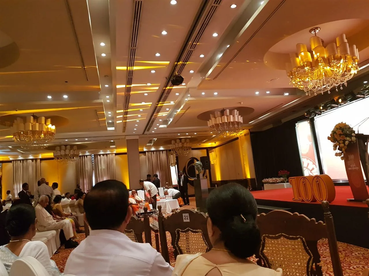 The image captures an indoor event, likely a formal celebration or ceremony. The setting features a large, elegantly decorated hall with a high ceiling adorned with large chandeliers. The lighting is warm, with yellow and orange tones, creating a welcoming and festive atmosphere. Several people are seated, facing a stage to the right, indicating a focus on a presentation or speech.
The stage itself is prominent, with a podium decorated with a bouquet of flowers and a large "100" displayed, suggesting a centennial celebration or similar milestone event. The audience appears engaged, seated on cushioned chairs, dressed in formal attire suitable for the occasion. The room's design includes ornate details and plush carpeting, enhancing the event's upscale ambiance. The scene reflects a gathering of attendees, possibly for a corporate or community milestone, with an emphasis on the ceremonial aspects of the event.
