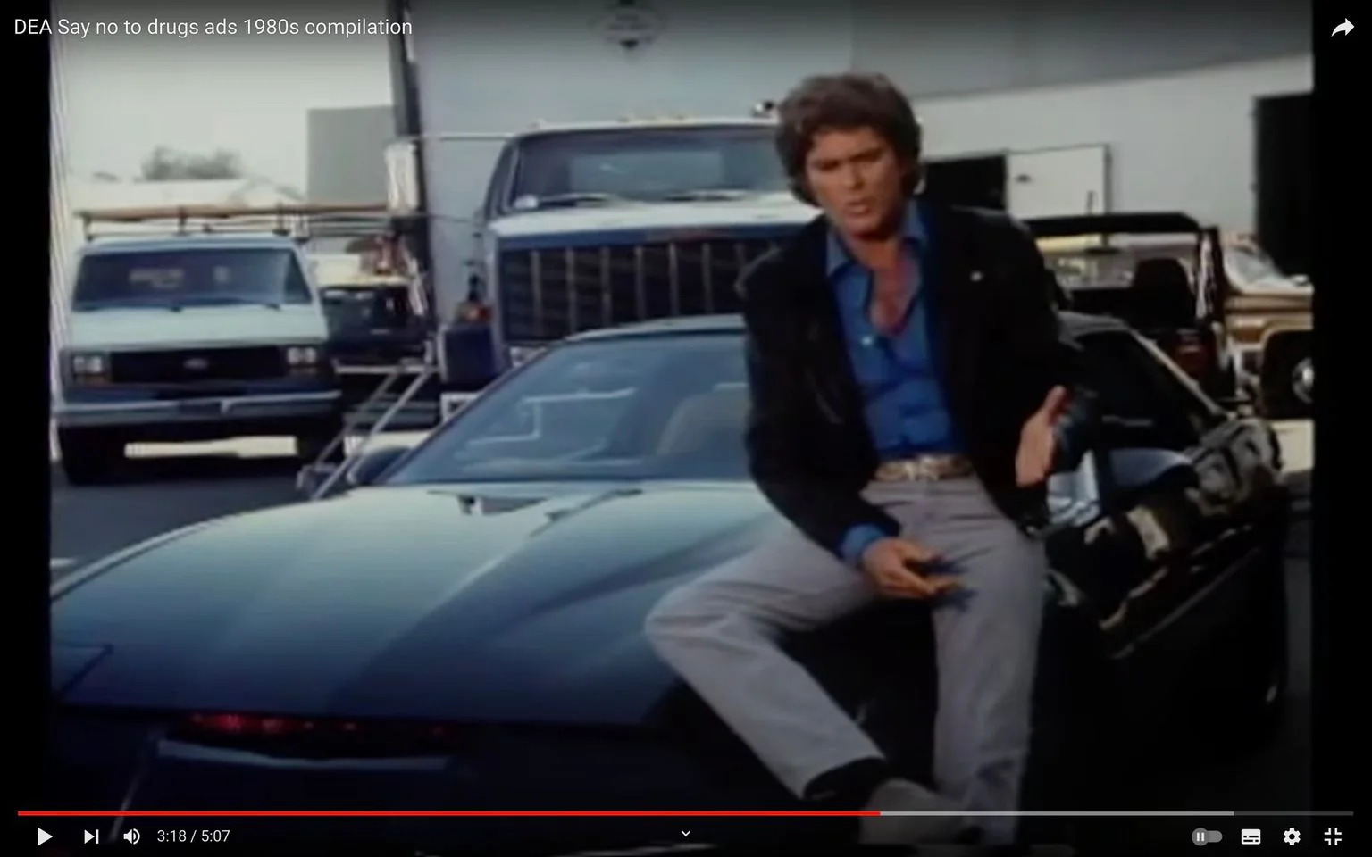 This image is a frame from a video compilation titled "DEA Say no to drugs ads 1980s compilation." The scene is set outdoors, likely on a studio lot or a similar location, as evidenced by the presence of several vehicles and trailers in the background. Prominently featured in the foreground is a person sitting on the hood of a sleek, black car. The individual is wearing a blue shirt, light-colored pants, and a black leather jacket, suggesting a casual yet stylish look typical of the 1980s. The car appears to be a classic model sports car, notable for its streamlined design and dark, shiny exterior.
The atmosphere of the scene is reminiscent of an anti-drug public service announcement, a popular campaign theme during that era. The setup includes a large truck and a van in the background, adding to the impression of an industrial or logistical setting. The lighting is natural, indicating that the scene was shot during the day. Despite the absence of captions or subtitles in this particular frame, the context strongly suggests the overall theme of promoting a drug-free lifestyle.