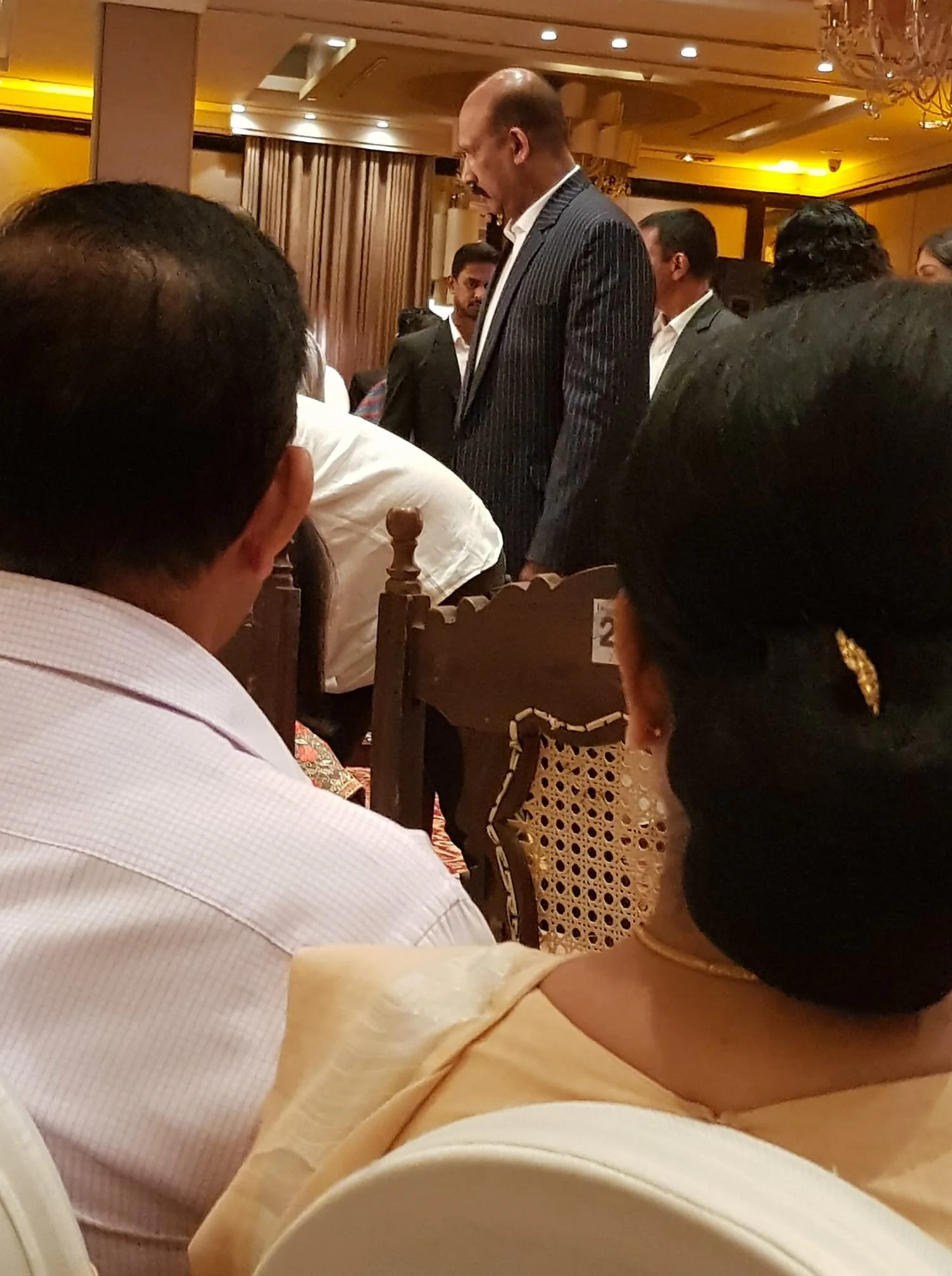 The image captures an indoor event with several people present. In the foreground, two individuals are seated with their backs to the camera; one is wearing a light-colored shirt and the other has neatly tied dark hair with a golden hairpin. The focus is on a man in a pinstripe suit standing in the center of the frame, identified as Kamal Gunaratne. He appears to be engaged in conversation, with a serious yet approachable demeanor. The setting is elegantly decorated, featuring soft lighting and chandeliers, suggesting a formal or celebratory occasion. The atmosphere is warm, with hues of gold and brown dominating the scene. Other attendees, dressed in formal attire, can be seen mingling in the background. The room has a sophisticated ambiance, enhanced by the ornate decor.