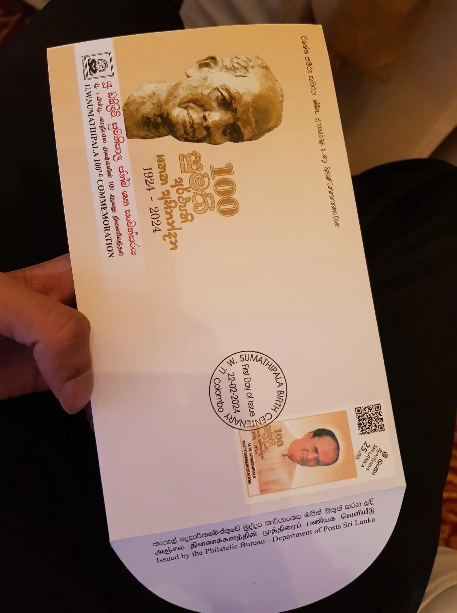 A special commemorative postal cover is depicted in the image, celebrating the 100th birth anniversary of U. W. Sumathipala. The cover features a bronze-toned bust of U. W. Sumathipala prominently displayed on the upper left side. Below it, the text "100th COMMEMORATION 1924 - 2024" is inscribed in a striking font. The event is marked as the "FIRST DAY OF ISSUE" on 22nd February 2024, in Colombo, as indicated by the circular stamp on the cover.
Adjacent to the stamp, there is a small photograph of U. W. Sumathipala, along with a printed QR code and stamp value. The cover is an official release by the Philatelic Bureau of the Department of Posts in Sri Lanka, highlighting its significance. The document is held in the hand of a person, suggesting it is being showcased at an event. The background is subtly blurred, focusing attention on the commemorative details of the envelope. The lighting is warm and soft, enhancing the formal and respectful tone of the image. This special issue signifies a tribute to the contributions of U. W. Sumathipala.