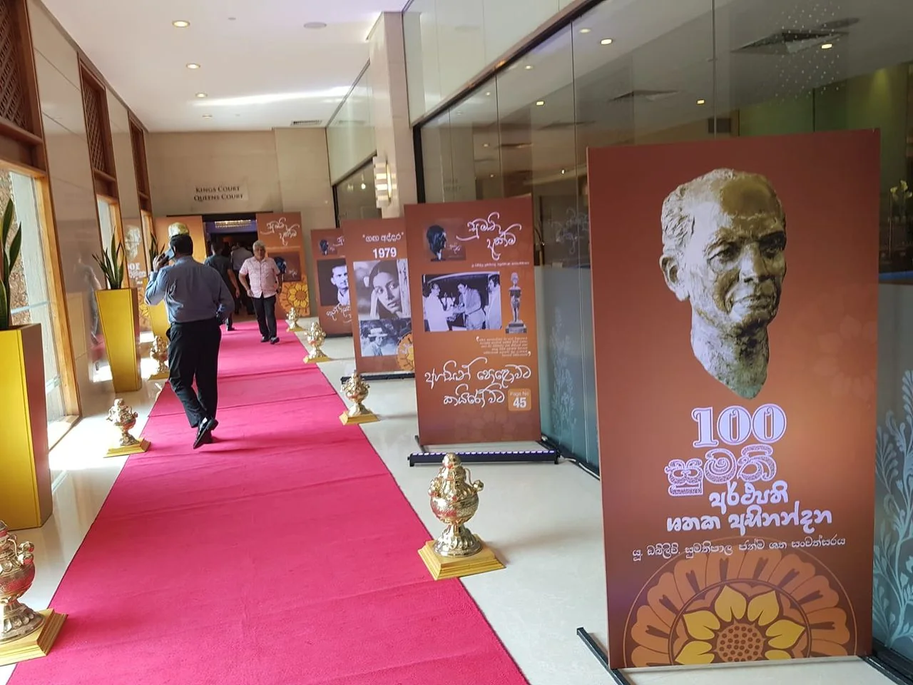 The image depicts an indoor event setting with a red carpet leading down a hallway. On either side of the carpet, there are several ornate urns placed on square stands. Along the right side, there are large posters mounted on stands, featuring images and text in a non-English script. The prominent poster on the right displays a bust of a man, with the number "100" prominently visible beneath it, suggesting a centennial celebration or commemoration. Another poster further down is marked with the year "1979," possibly indicating a historical event or milestone.
The walls of the hallway are lined with glass panels and decorative wooden partitions. The lighting is bright and evenly distributed, creating a formal and inviting atmosphere. Several individuals are walking along the carpet, dressed in a mix of casual and business attire, suggesting a blend of formal and informal participation. The setting appears to be a formal reception or exhibition, possibly honoring a significant figure or event. The overall mood is respectful and commemorative, with a focus on celebrating historical or cultural heritage.