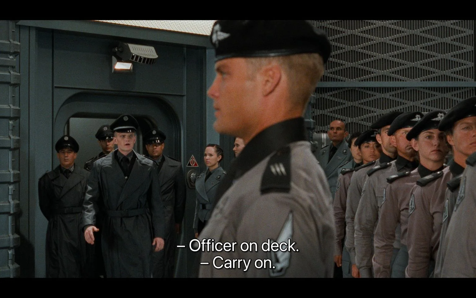In this scene from the film "Starship Troopers," a group of military personnel is seen standing in uniform formation inside what appears to be a spacecraft or military facility. The setting features a metallic, industrial interior with a textured, grid-like wall in the background. The personnel are wearing gray and black uniforms with matching caps, which suggest a hierarchical military structure.
Neil Patrick Harris is among the characters present in the scene. He is dressed in a distinctive black uniform and is positioned centrally, walking confidently with an authoritative presence. The other characters, standing in neat rows, appear alert and disciplined, reflecting a formal military environment. The image is captured with a focus on order and discipline, indicative of a structured and hierarchical setting.
The lighting is bright and functional, emphasizing the utilitarian design of the military setting. Subtitles at the bottom of the image read, "– Officer on deck." and "– Carry on." These lines suggest a moment of acknowledgment and command within the scene, reinforcing the hierarchical nature of the military environment depicted.