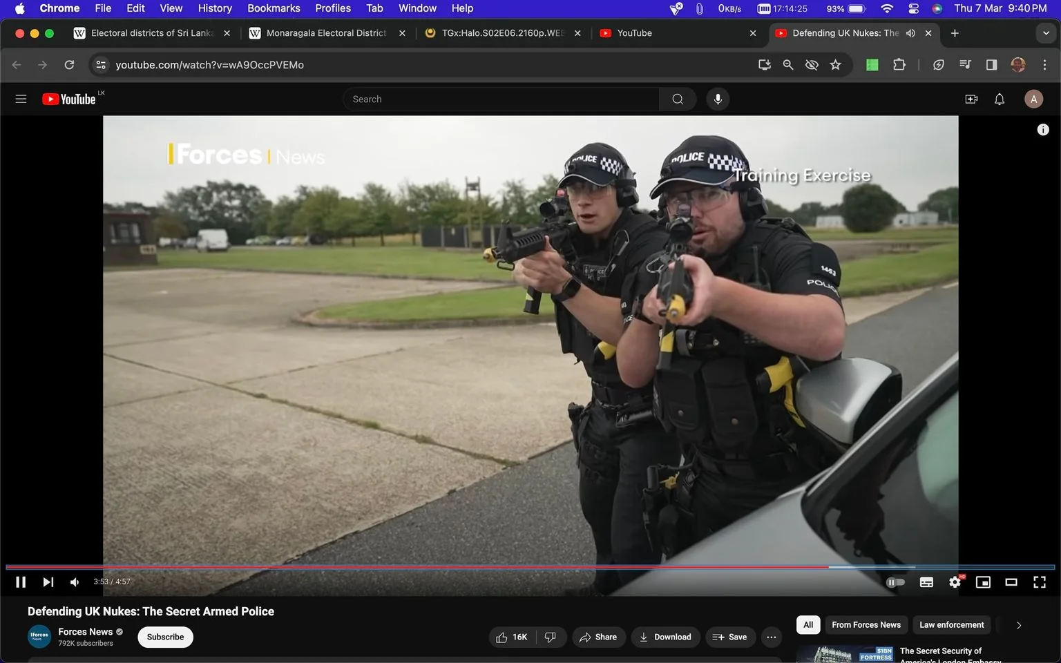 The image is a screenshot from a YouTube video titled "Defending UK Nukes: The Secret Armed Police," published by Forces News. The scene depicts an outdoor training exercise involving two police officers, both equipped with tactical gear and firearms. They are positioned beside a vehicle, with their weapons aimed forward, indicating a simulation of an armed confrontation. Both officers wear black uniforms, caps with a checkered pattern, and protective eyewear, emphasizing their readiness and focus. The setting appears to be an open, grassy area with a few buildings and parked vehicles in the background, suggesting a controlled environment for training purposes. The mood is tense and serious, reflecting the gravity of their simulated task. The screenshot also includes the Forces News logo and the phrase "Training Exercise" overlaying the image, enhancing the context of the activity.