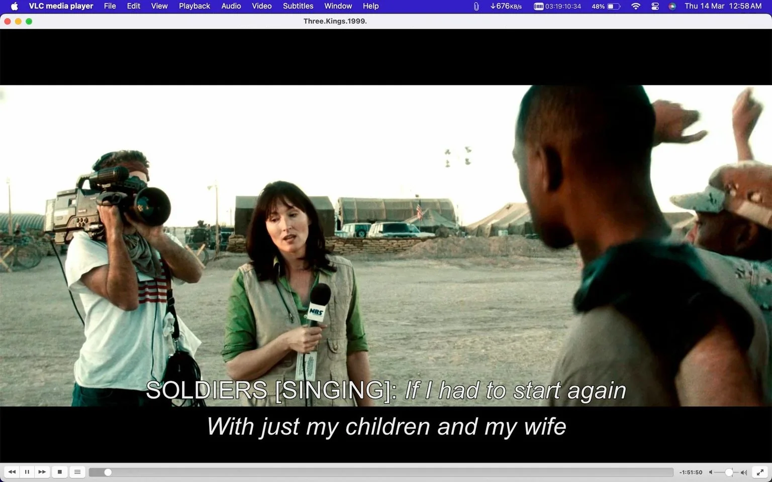 In this video frame from the movie "Three Kings," the setting is an outdoor desert environment, likely a military camp, visible from the sandbags and tents in the background. The image captures a scene where a news reporter is conducting an interview. The reporter holds a microphone labeled "NBS" and stands next to a cameraman who is filming the event. The reporter appears focused and composed, dressed in a green shirt and a beige vest, matching the gritty, muted tones of the setting. The cameraman, dressed casually, is capturing the action using a large, professional video camera.
In the foreground, a soldier is partially visible, facing the reporter. The mood suggests a mix of seriousness and casualness, typical of a field report amidst a military setting. Overlaid at the bottom of the image are subtitles of soldiers singing: "If I had to start again / With just my children and my wife." The video frame is displayed on the VLC media player interface, indicating the playback environment on a computer screen. The lighting is natural, suggesting it is either early morning or late afternoon.