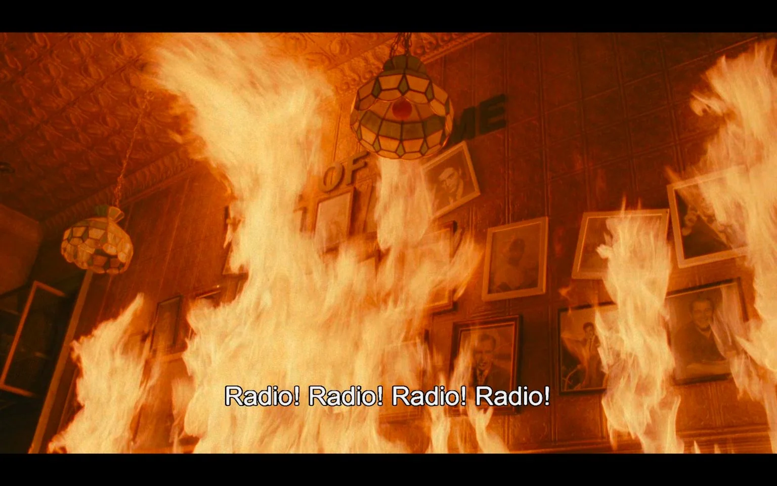 The image is a dramatic scene from the film "Do the Right Thing." It depicts an intense fire engulfing a wall adorned with framed photographs. The flames are vivid, casting an orange glow over the scene, creating a sense of chaos and urgency. The intricately patterned ceiling and hanging lamp suggest the setting is an indoor space, possibly a restaurant or community center, given the "OF ME" text partially visible on the wall, which might be part of a larger phrase like "Hall of Fame."
The scene is filled with energy and tension, emphasized by the repetitive caption "Radio! Radio! Radio! Radio!" suggesting a climactic moment involving sound or communication. The composition focuses on the destructive power of the fire, with the photographs serving as poignant reminders of personal or community history being consumed by the flames. This visual effectively conveys themes of conflict and change central to the film's narrative.
- Captions / subtitles: "Radio! Radio! Radio! Radio!"