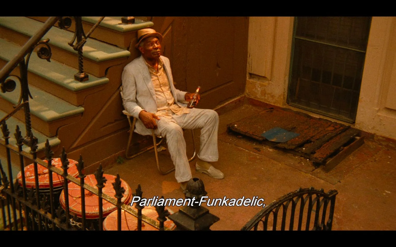 The image is a scene from the film "Do the Right Thing." It shows a man sitting outdoors on a folding chair near a stoop in an urban setting. He is dressed in a light gray suit with a matching hat, and he's holding a bottle, possibly a drink, in his right hand. His expression appears relaxed and contemplative. The setting features worn stairs leading to a building entrance and several red trash cans lined up near a black iron fence. The lighting suggests it is daytime, with warm tones casting a soft glow across the scene, which complements the earthy colors of the building and the man's attire. The atmosphere conveys a laid-back, everyday moment on a city street. The subtitle reads "Parliament-Funkadelic," adding an auditory element to the visual scene.
- Captions / subtitles: "Parliament-Funkadelic,"