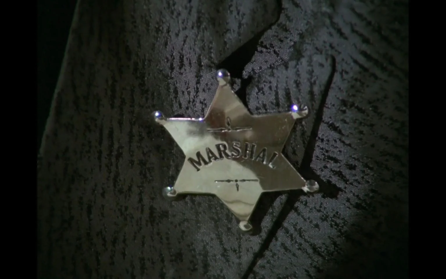 The image is a close-up shot of a shiny, silver star-shaped badge with the word "MARSHAL" engraved on it. This badge stands out against a textured, dark-colored fabric, likely part of a uniform or costume. The lighting highlights the metallic surface of the badge, creating reflections that draw attention to the engraved text. The scene is linked to "Living in Harmony," which suggests a context related to Western or law enforcement themes. The badge's design is simple yet iconic, reminiscent of traditional law enforcement insignia often seen in Western films or television shows. The overall mood is serious and authoritative, emphasizing the role of a marshal or law figure. The fabric in the background features a subtle pattern, adding depth and texture to the image.