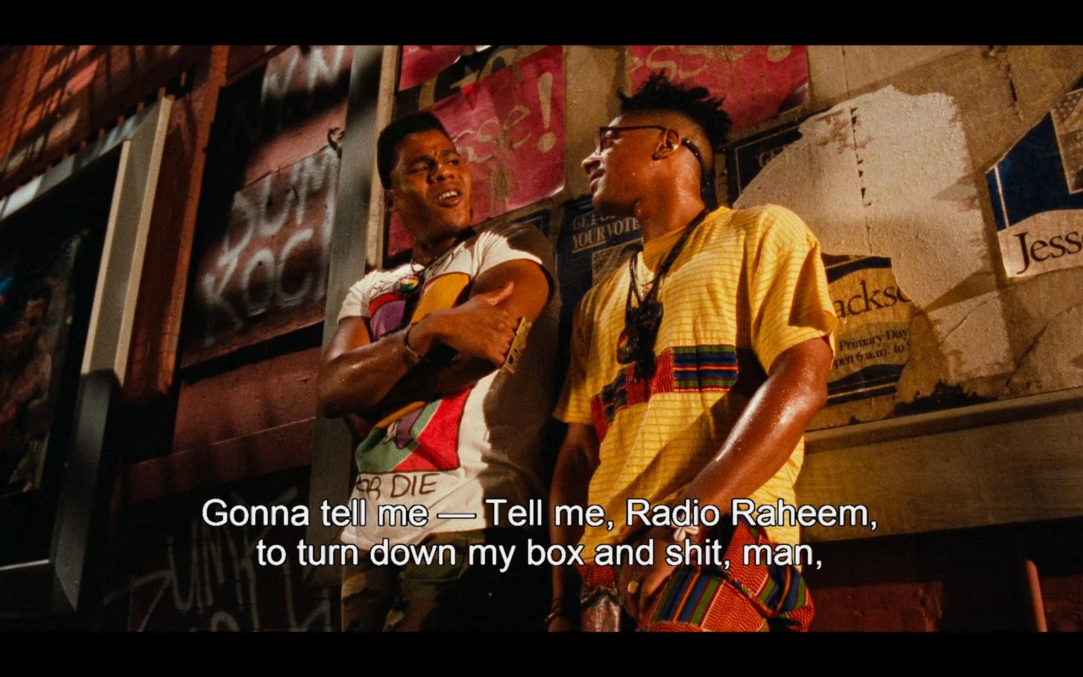 In this scene from "Do the Right Thing," two characters are engaged in an animated conversation on a dimly lit street at night. The character on the left is wearing a white t-shirt with colorful graphics, standing with arms crossed and a defiant expression. The character on the right wears a yellow, striped shirt with a vibrant pattern, accessorized with glasses and jewelry. Their posture suggests a casual camaraderie, although the conversation seems to carry some tension or urgency.
The background is adorned with a variety of posters, some of which are partially torn or covered with graffiti, contributing to the gritty urban atmosphere typical of the film. The lighting is warm, casting soft shadows and enhancing the vibrancy of their clothing, while the wall behind them is a mix of brick and plaster, adding texture to the scene.
The subtitle reads, "Gonna tell me — Tell me, Radio Raheem, to turn down my box and shit, man," indicating a conversation possibly about music or personal space. The use of "Radio Raheem" is a reference to a character known for his love of loud music, a thematic element in "Do the Right Thing." The scene captures the essence of street life and the dynamics within the community depicted in the movie.
