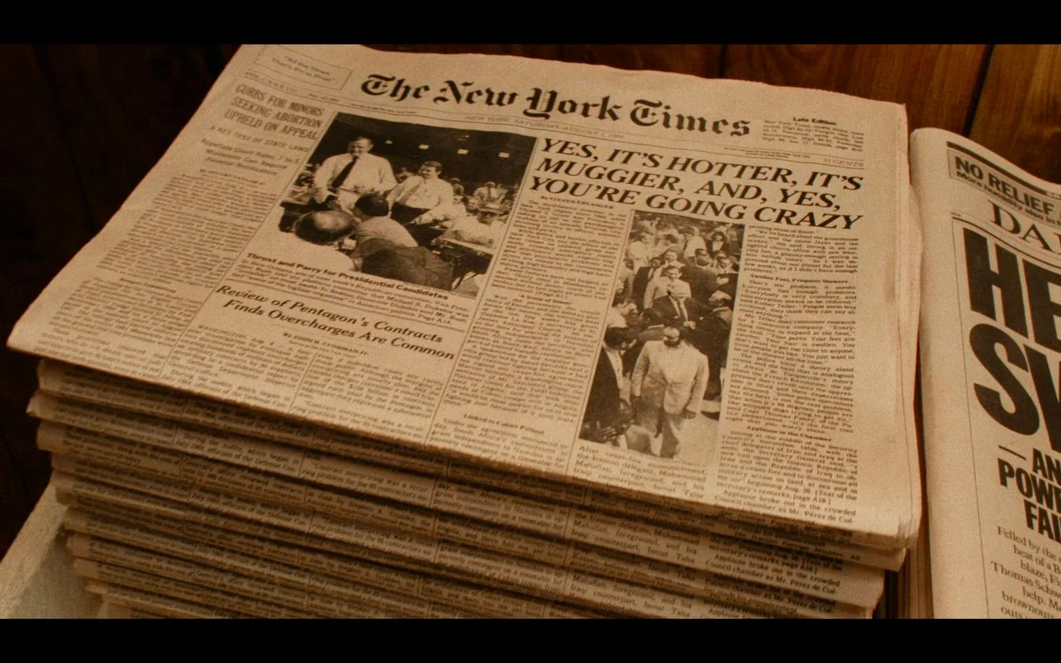 The image depicts a stack of newspapers with a prominent headline from "The New York Times." The visible headline reads, "YES, IT'S HOTTER, IT'S MUGGIER, AND, YES, YOU'RE GOING CRAZY," capturing the oppressive heat theme from the film "Do the Right Thing." The newspapers are neatly piled, suggesting a setting inside a shop or newsstand. The overall color palette is a muted sepia, evoking a vintage or nostalgic feel, which aligns with the film’s late 1980s aesthetic.
The top newspaper features a black and white photograph of people gathered, possibly reflecting the community scenes from the movie. To the left, an article titled "Review of Pentagon's Contracts Finds Overcharges Are Common" is visible, adding a political and serious tone to the front page. The name "The New York Times" is prominently displayed at the top along with the date, reinforcing the credibility and authenticity of the setting. This newspaper piece is a key element in establishing the film’s setting during a particularly hot day, a central element around which the movie's plot unfolds. The image effectively captures a snapshot of the social and environmental conditions central to "Do the Right Thing."