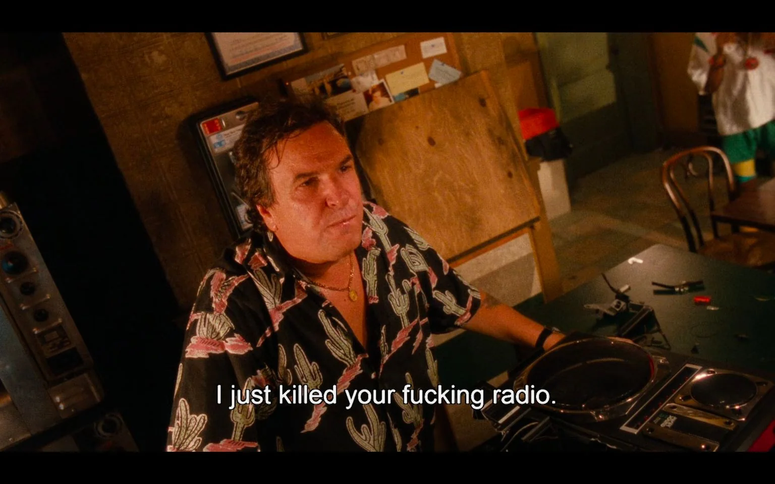 The screenshot is from the film "Do the Right Thing." It depicts a scene set indoors, likely in a shop or common area. The lighting is warm and somewhat dim, contributing to an intense and confrontational atmosphere. In the foreground, a man is standing behind a table with various electronic components scattered around, including a radio. He appears to be speaking forcefully, as indicated by the subtitle: "I just killed your fucking radio." His expression is serious, suggesting a moment of tension or conflict. He is wearing a black shirt adorned with cactus patterns, a necklace, and has an earring, adding to the character's distinctive style. The background shows a bulletin board and part of a vending machine. This scene captures an emotionally charged interaction, reflective of the film's exploration of social and personal conflicts.
- Captions / subtitles: "I just killed your fucking radio."