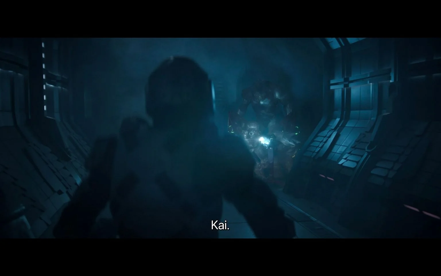 The image is a screenshot from the show "Halo," depicting a tense scene in a dimly lit futuristic corridor. In the foreground, a figure in a suit, presumably of armor, is seen with their back to the viewer, facing a large, imposing creature or figure in the distance. The corridor is metallic, with intricate paneling and a low, moody blue light creating a sense of suspense and danger. The figure in the background appears to be of significant size, adding to the tension of the moment. The environment suggests a high-tech setting, typical of a sci-fi narrative. The subtitle “Kai.” is visible at the bottom of the image, likely indicating a character named Kai is part of the scene. The overall atmosphere is dark and foreboding, contributing to the dramatic tension in the scene.