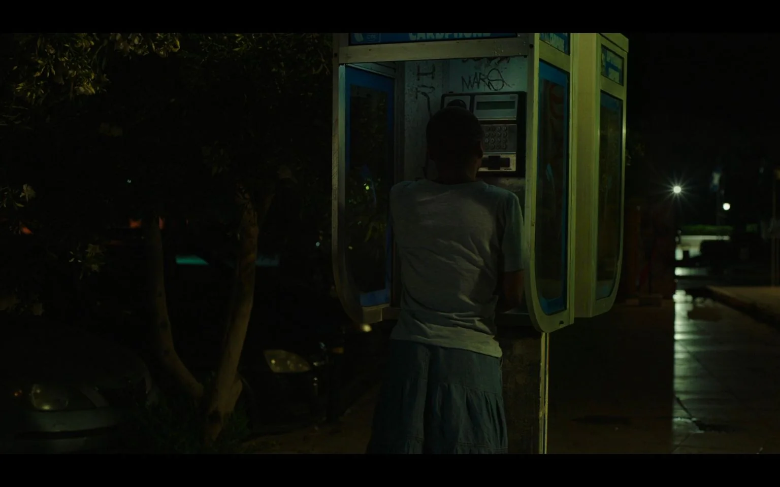 The image depicts a nighttime scene centered around a person standing at a phone booth. The street is dimly lit, with the glow of streetlights reflecting off the wet pavement, suggesting a recent rain. The person, whose back is facing the camera, is wearing a light gray t-shirt and a denim skirt. Their posture is slightly leaned forward as they engage with the payphone inside the booth. The booth itself is illuminated, casting a soft blue and white light that contrasts with the surrounding darkness. Graffiti is visible on the phone box, with the word "MAKS" discernible. The background includes parked cars and silhouetted trees, adding to the urban setting's quiet ambiance. The overall mood is solitary and introspective, highlighted by the subdued lighting and empty street.