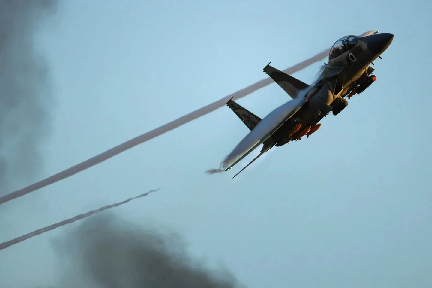 The image depicts a military jet, likely a fighter aircraft, captured mid-flight against a clear blue sky. The jet is angled sharply, suggesting a maneuver or ascent, with vapor trails streaming from its wings. The lighting emphasizes the sleek design of the aircraft, highlighting its aerodynamic shape against the soft hues of the evening or morning sky. The aircraft's surface has a reflective sheen, and its markings and insignias can be seen, suggesting identification with a specific air force or military unit. The overall scene conveys a sense of speed and precision, characteristic of modern aerial combat aircraft. The background reveals faint, dissipating smoke trails, indicating recent activity or maneuvers. The image is dynamic, capturing the essence of modern military aviation technology.