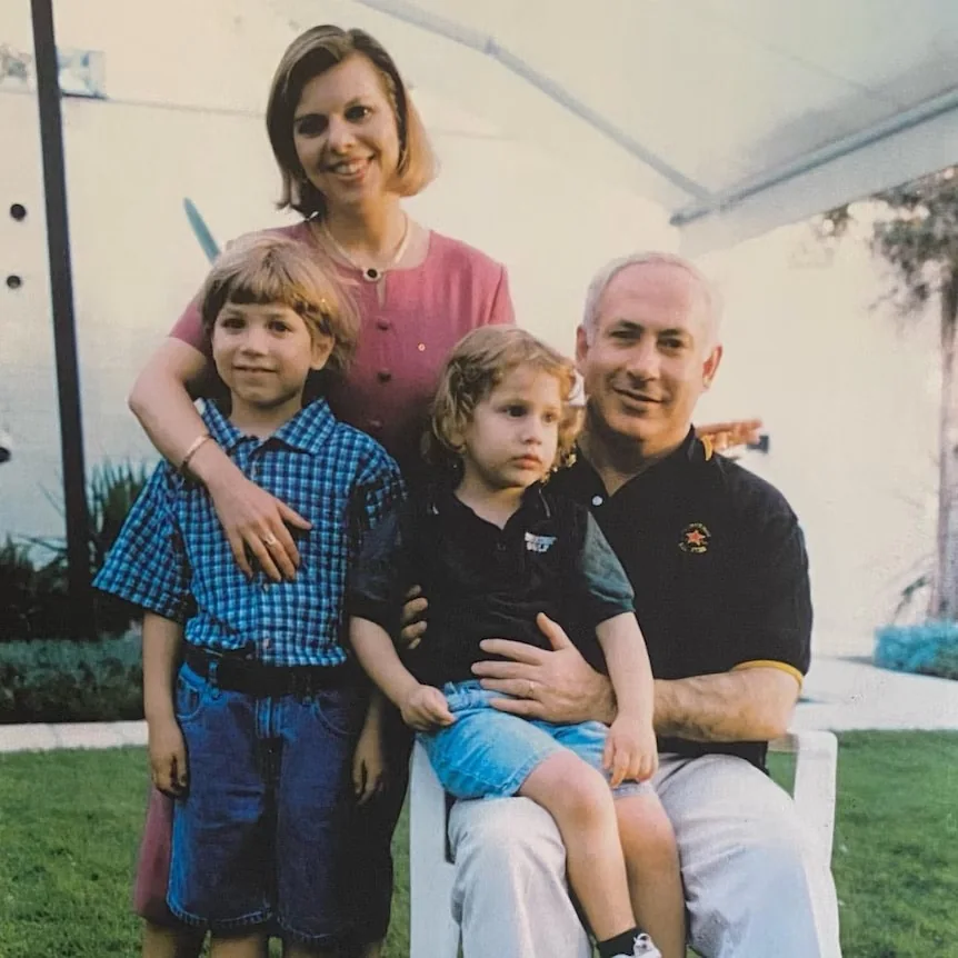 The image features a family portrait set outdoors during the daytime. In the foreground, a man is seated on a white chair, holding a young child on his lap. The child appears to be around toddler age, wearing a black shirt and light blue shorts. Alongside them, a slightly older child stands, wearing a checkered blue shirt and dark shorts, with a gentle smile. Behind them, a woman stands, dressed in a pink dress, with one arm around the older child, projecting a warm and caring demeanor.
The background suggests a garden-like setting, with greenery and a partially visible structure, possibly a canopy or tent, adding depth to the scene. The lighting is bright and natural, indicating a sunny day. The overall mood of the photograph conveys familial warmth and closeness. The image likely captures a moment of family bonding and leisure. Notably, the hint identifies the individuals as Benjamin, Sarah, Yair, and Noa Netanyahu, with a specific mention of Noa Netanyahu within the context of this family group.