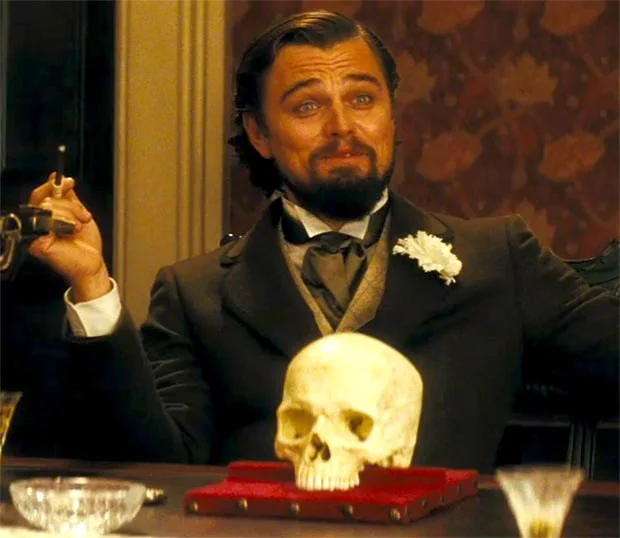 Monsieur Calvin Candie, played by Leo DiCaprio, is depicted in an opulent interior scene from the film "Django Unchained." He sits confidently at a table adorned with a human skull placed on a red velvet cushion, which adds a sinister and dramatic touch to the setting. Candie is dressed in a formal, dark suit with a vest, a white shirt, and a cravat, exuding an air of wealth and authority. A white flower is pinned to his lapel, enhancing his sophisticated appearance. His expression is a mix of amusement and smugness, suggesting a moment of triumph or condescension. The room's warm lighting casts a glow on his face, highlighting his expressions and enhancing the atmosphere's intensity. The background shows rich wallpaper and elegant decor, contributing to the scene's luxurious yet tense mood. The overall composition captures a pivotal and intense moment, reflecting the character's complex personality.