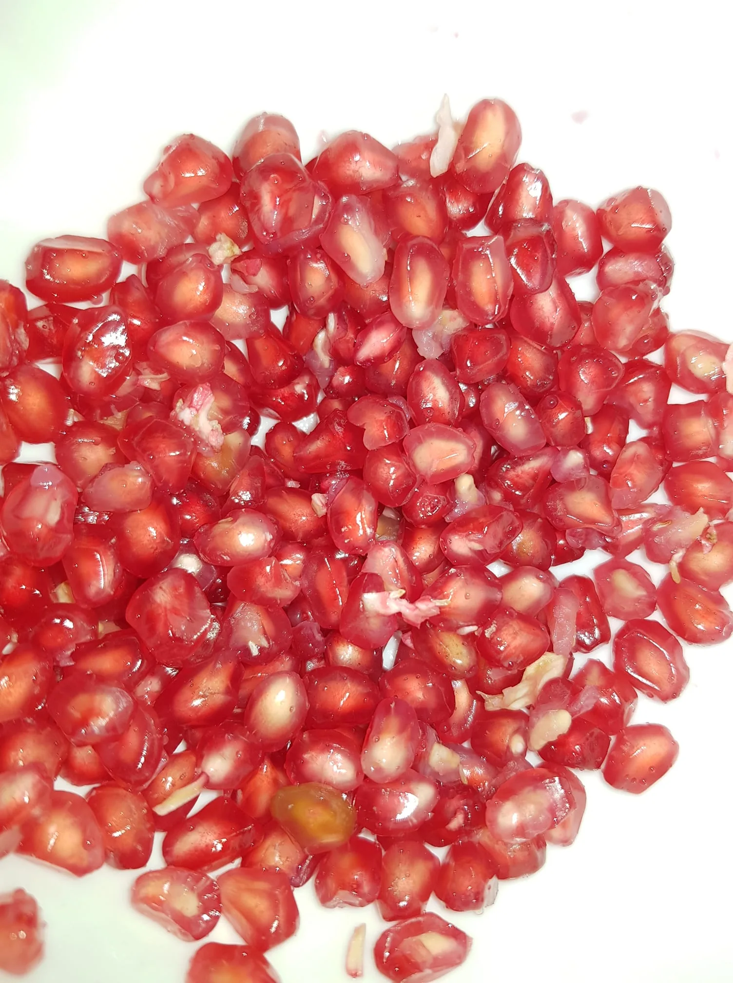 The image shows a close-up of pomegranate seeds, also known as arils, scattered on a white surface. These seeds are vibrant red, glossy, and translucent, indicating they are fresh and juicy. The lighting highlights the seeds' shiny texture, enhancing their appealing appearance. Some white bits of the pith are visible among the seeds, which is common when they are freshly extracted from the fruit. The background is plain, focusing attention entirely on the pomegranate seeds. The image captures the essence of fresh produce and could be used in contexts related to healthy eating or culinary arts.