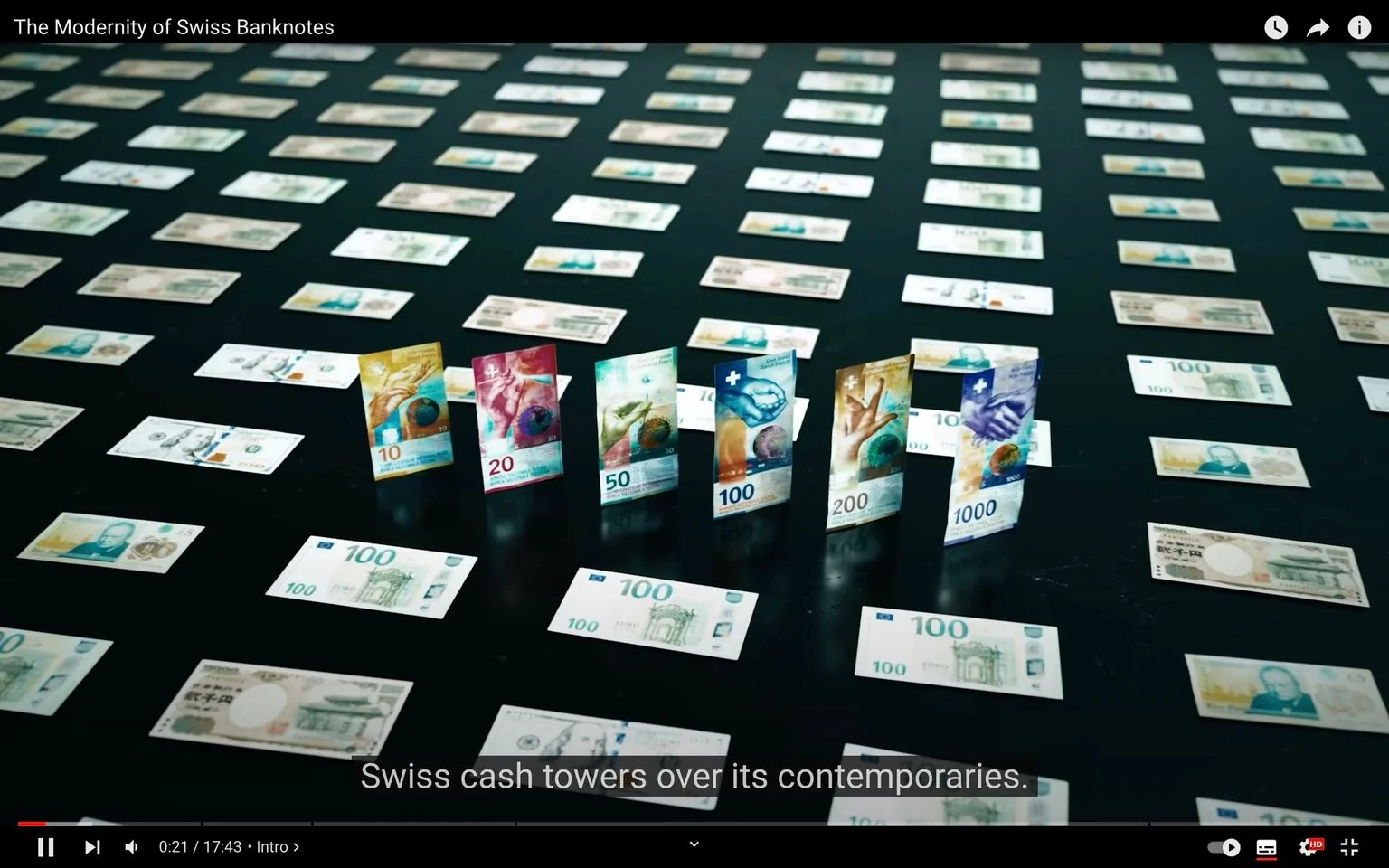 The video frame showcases an organized display of Swiss banknotes, with several bills standing upright in the foreground. The title "The Modernity of Swiss Banknotes" is visible at the top, suggesting a focus on the innovative design of these notes. In the center, vertically positioned notes in denominations of 10, 20, 50, 100, 200, and 1000 Swiss francs are prominently featured. These notes are vibrant, with colorful designs that highlight modern aesthetic elements, such as abstract representations and images of hands holding globes. The background features older, flat banknotes, arranged in neat rows across a dark surface, creating a contrast with the modern designs. The subtitle "Swiss cash towers over its contemporaries." emphasizes the advanced design qualities of Swiss currency. The overall composition, with the contemporary notes standing out, reflects a theme of progression and innovation in currency design. The lighting is soft and evenly distributed, enhancing the clarity and color of the banknotes.