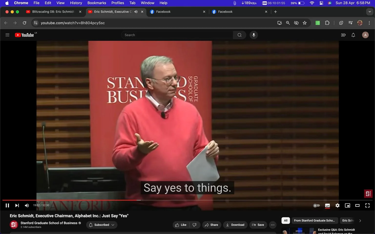 Eric Schmidt is captured mid-presentation in a lecture hall, highlighted against a backdrop featuring a red banner with the words "STANFORD BUSINESS GRADUATE SCHOOL." The setting is clearly academic, with wooden paneling suggesting a formal environment. Eric Schmidt is wearing a bright pink sweater over a collared shirt, holding a sheet of paper, and gesturing with his hand, emphasizing a point to his audience. The lighting is warm and focused on him, indicating he is the center of attention in this scene. His demeanor is engaged and confident, likely reflecting the motivational nature of his talk.
The YouTube video title is "Eric Schmidt, Executive Chairman, Alphabet Inc.: Just Say 'Yes'," and it is hosted on the Stanford Graduate School of Business channel. The subtitle "Say yes to things." appears at the bottom, capturing a key takeaway from his speech. The interface shows a Chrome browser window, with visible tabs at the top and typical YouTube controls below the video.