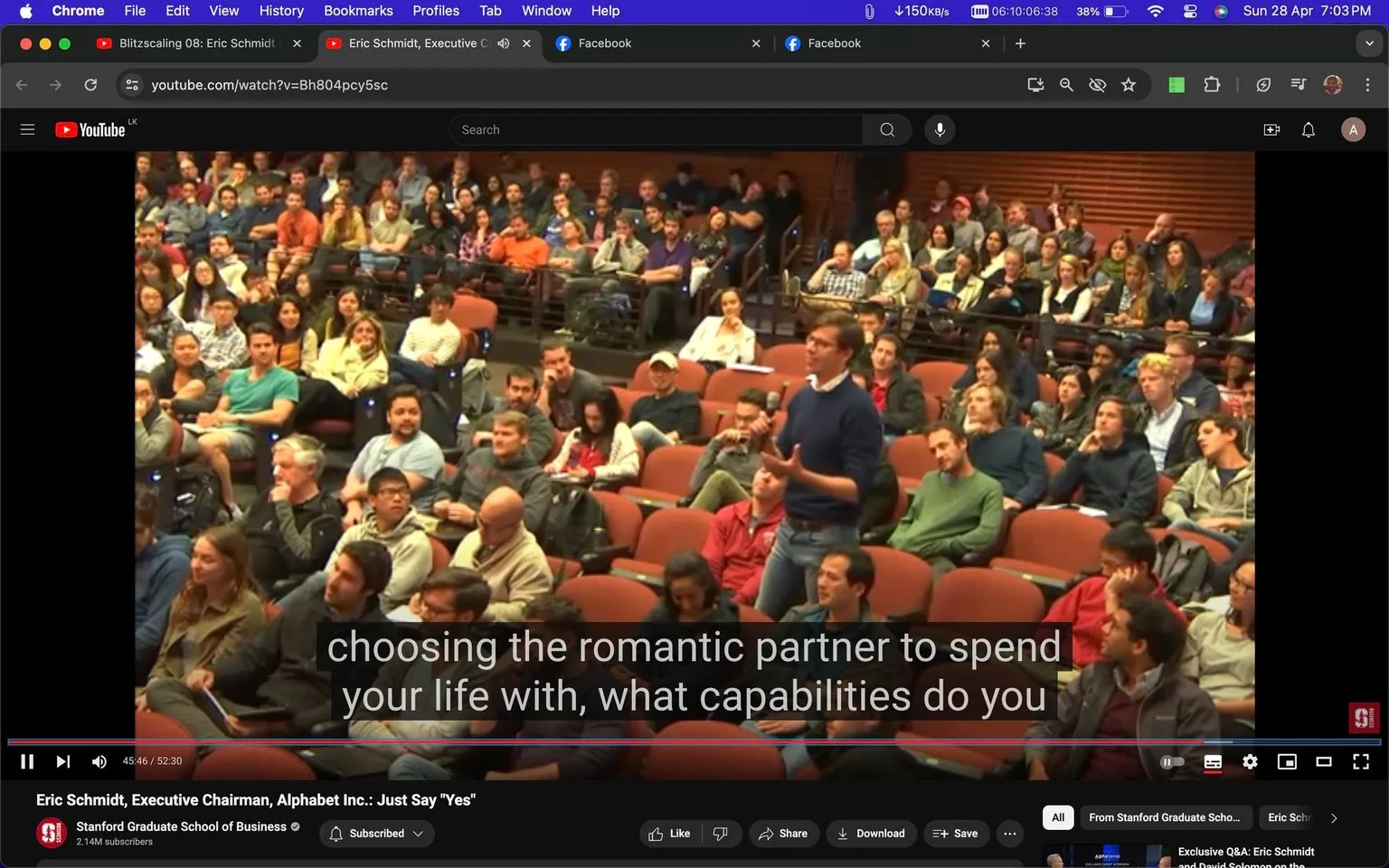 1) The image is a screenshot from a YouTube video titled "Eric Schmidt, Executive Chairman, Alphabet Inc.: Just Say 'Yes'" from the Stanford Graduate School of Business channel. The scene is set in a large lecture hall filled with an audience seated on red chairs. A man, standing in the aisle, appears to be asking a question. He is wearing a blue sweater and glasses, holding a microphone, and the audience is attentively listening. The event seems to be a lecture or Q&A session, with a focus on business or technology topics. The background is filled with people of diverse ages and attire, suggesting a university setting. The lighting is bright, highlighting the engaged expressions of the attendees. Captions at the bottom of the screen capture a portion of a question being asked.
2) On-screen text block:
- Title: "Eric Schmidt, Executive Chairman, Alphabet Inc.: Just Say 'Yes'"
- Channel / profile: "Stanford Graduate School of Business"
- Site / app: "YouTube"
- Captions / subtitles: "choosing the romantic partner to spend your life with, what capabilities do you"