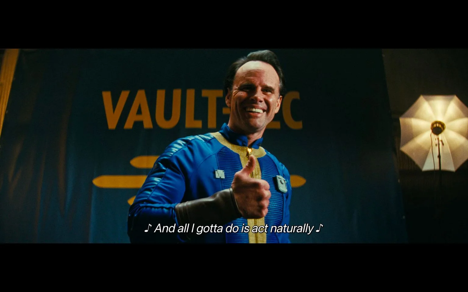 The image is a screenshot from the "Fallout TV show." It features a person wearing a distinctive blue jumpsuit with yellow accents, typical of the "Vault" attire from the Fallout universe. The person is smiling and giving a thumbs-up gesture, exuding a confident and cheerful demeanor. The backdrop prominently displays the word "VAULTEC," connecting to the Vault-Tec Corporation from the Fallout series, known for its role in the post-apocalyptic setting of the franchise.
A lighting setup, including a softbox light on a stand, is visible, suggesting the scene is part of a media or promotional event. The warm lighting enhances the optimistic and lively atmosphere. The caption reads, "♪ And all I gotta do is act naturally ♪," indicating a lighthearted or musical moment in the show. The composition centers on the character, capturing a moment of engagement with the audience, aligning with the show's themes of survival and adaptability in a post-apocalyptic world.