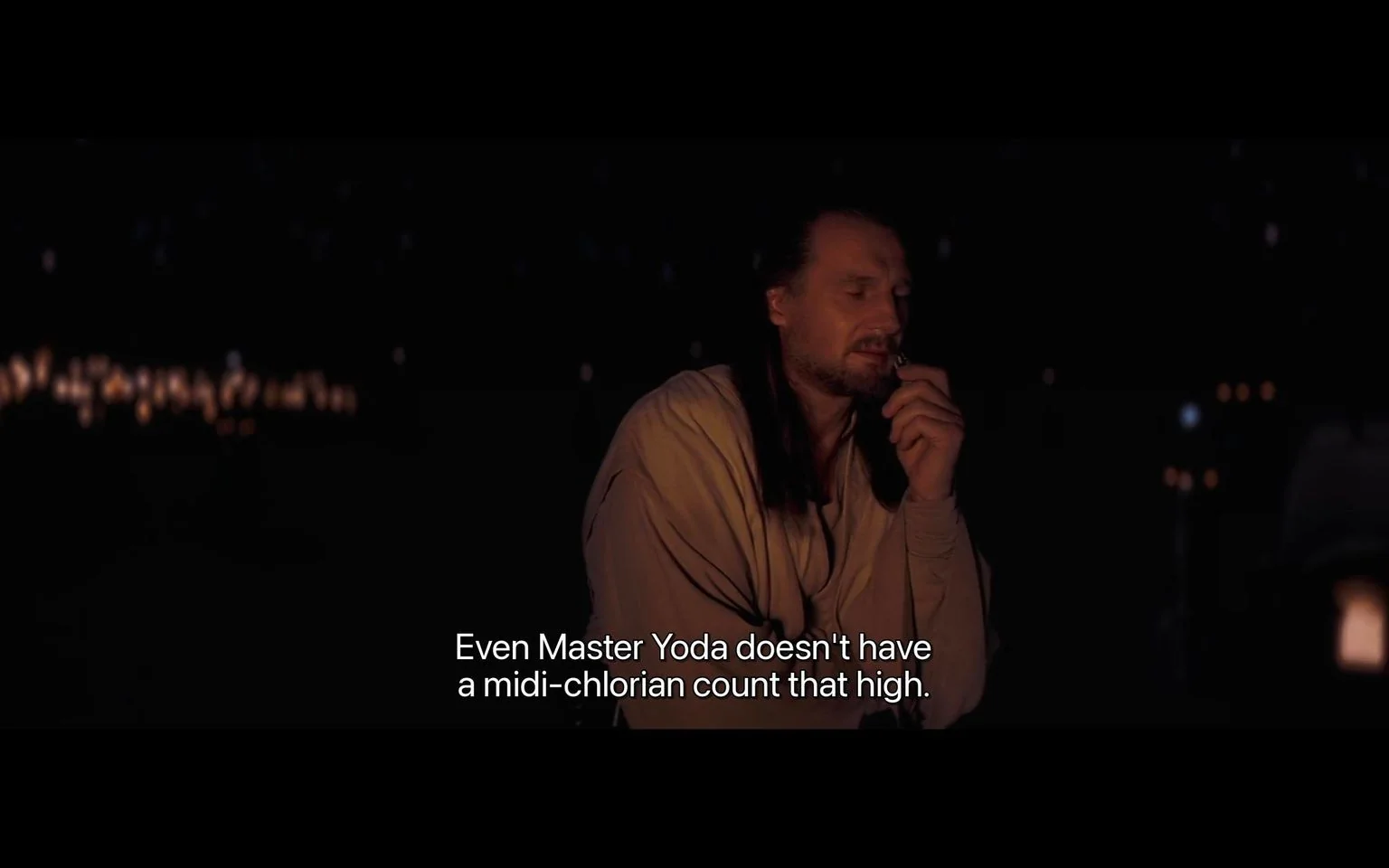 The image is a night-time scene from a "Star Wars" film. It features a character in a contemplative pose, partially illuminated by what appears to be a campfire or similar light source, giving the setting a warm, ambient glow amidst the darkness. The character is dressed in traditional Jedi robes, with long hair and a thoughtful expression, suggesting a moment of reflection or discussion. In the background, small points of light suggest a distant cityscape or stars, adding depth to the scene. The dialogue at the bottom reads, "Even Master Yoda doesn't have a midi-chlorian count that high," indicating a conversation about the Force and its connection to midi-chlorians, a key concept in the "Star Wars" universe. The mood is serious and introspective, fitting the themes of destiny and power that are often explored in the series. This frame captures a moment of revelation or significance within the story.