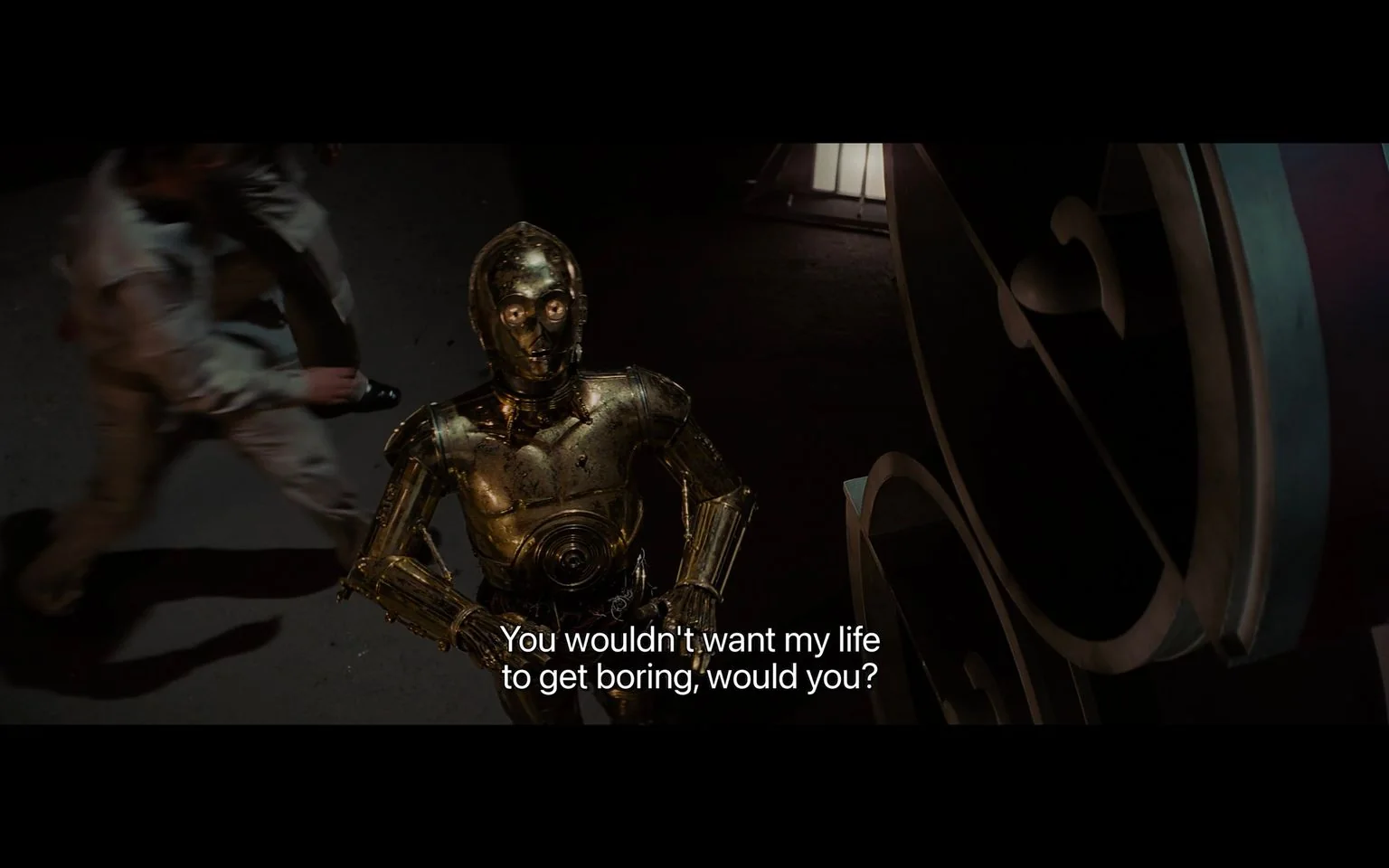1) Rich description: The image is a screenshot from a scene in "Star Wars." It features the iconic character C-3PO, a humanoid robot with a shiny golden exterior, standing indoors. The robot's design is detailed, with visible joints and panels that reflect the light, enhancing its metallic appearance. C-3PO is looking up, possibly engaging with someone off-screen, and is positioned in the center foreground. The background is dark, with a slightly blurred figure moving past, adding a sense of motion and urgency to the scene. The lighting is soft yet dramatic, highlighting the robot's features against the shadowy setting. The subtitle, "You wouldn't want my life to get boring, would you?" suggests a whimsical or humorous tone in C-3PO's dialogue. The environment appears industrial, with structures that have an angular and mechanical aesthetic, consistent with the "Star Wars" universe's futuristic theme.
2) On-screen text block:
- Captions / subtitles: "You wouldn't want my life to get boring, would you?"