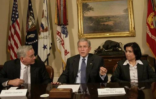 The image depicts a formal meeting setting with three individuals seated at a conference table. The central figure is speaking and gesturing with his hands, suggesting a discussion or presentation. To his left, a nameplate reads "Walter Isaacson," identifying the man seated and listening attentively. To the speaker's right, another nameplate identifies "Minister Tzipi Livni," who is also paying close attention.
The room is adorned with multiple flags in the background, adding to the official and formal atmosphere. There is a framed painting and a decorative sculpture on a table behind them, enhancing the dignified setting. All three individuals are dressed in formal business attire, with suits and ties, emphasizing the seriousness of the meeting.
The lighting is bright and even, highlighting the expressions and details of the participants. The focus is on the central figure, suggesting he is leading the discussion. The overall mood is professional and composed, indicative of a high-level political or diplomatic dialogue.