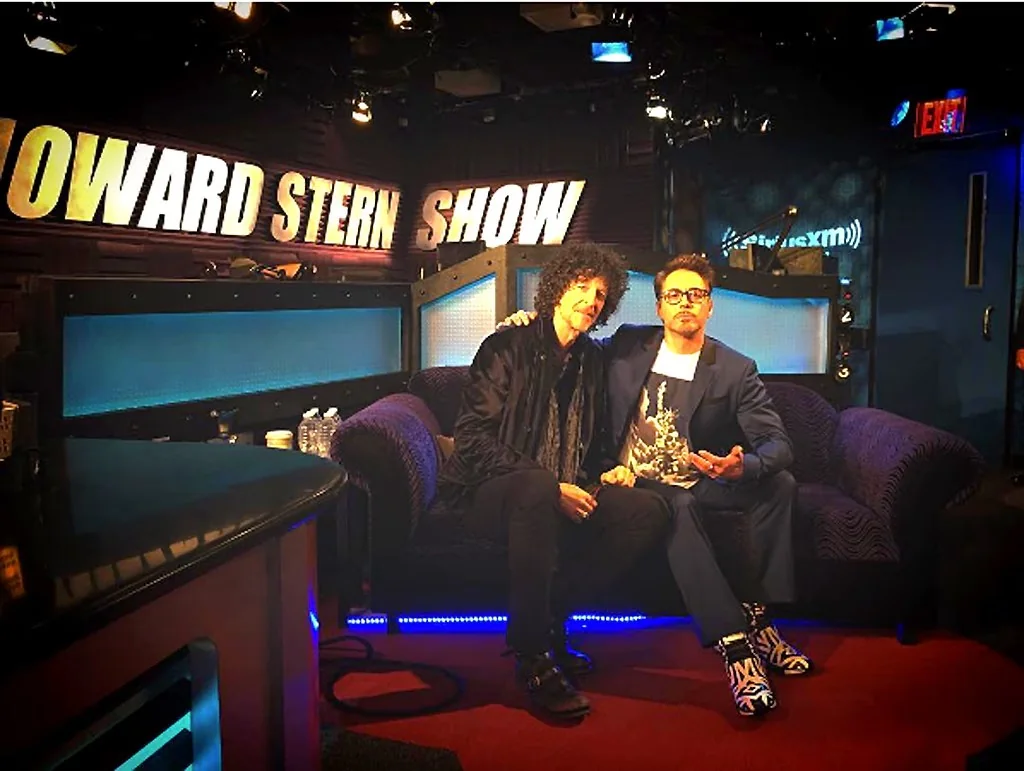 The image shows the interior of a radio studio where Howard Stern is seated on a couch next to a guest. The setting is the "Howard Stern Show," as indicated by the prominent sign in the background glowing with bright yellow letters. The studio is dimly lit, creating a cozy atmosphere, with the couch and surrounding decor contributing to this ambiance. Howard Stern, noted for his distinctive curly hair, is casually dressed in dark attire, leaning slightly toward his guest with a friendly demeanor. The guest, dressed in a sharp suit, strikes a relaxed pose, his expression suggesting engagement with the conversation. The studio's aesthetic is modern, with blue and purple hues from the lighting adding a vibrant touch. The SiriusXM logo can be seen in the background, indicating the network broadcasting the show. Bottled water and coffee cups are visible on a side table, suggesting a typical studio setup for guests. The overall mood is professional yet relaxed, fitting for an interview setting.
- Title: "Howard Stern Show"
- Site / app: "SiriusXM"