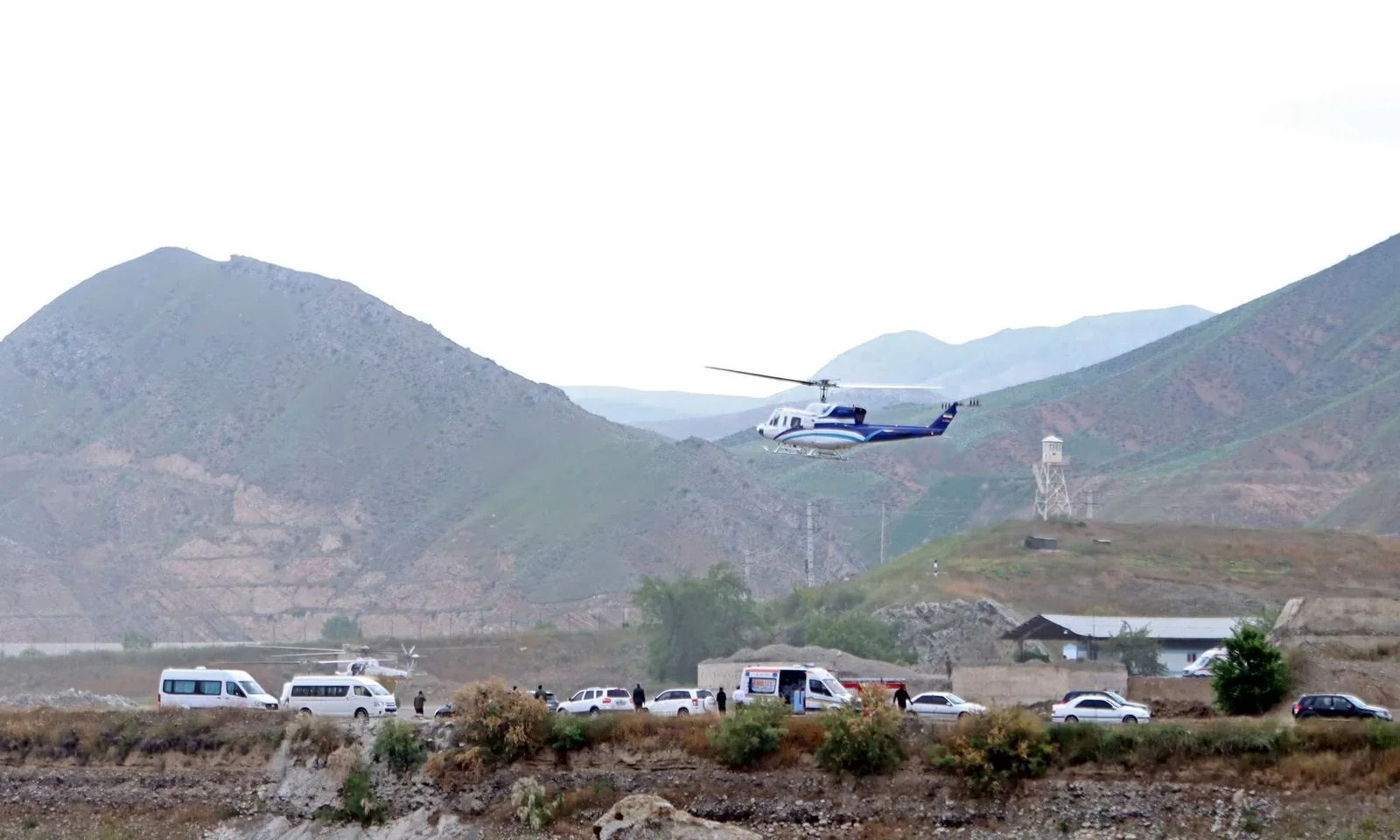 1) Rich description: The image depicts an outdoor scene with a helicopter in mid-air, flying over a landscape of rolling hills and mountains. In the foreground, there are several white vehicles, including vans and cars, parked on a dirt road. The helicopter is painted in blue and white, contrasting against the muted greens and browns of the mountainous backdrop. A group of people can be seen near the vehicles, possibly engaged in some coordinated activity. The lighting is natural, suggesting daytime, though the sky appears overcast, casting a diffuse light over the scene. A watchtower is visible in the distance, adding an element of security or surveillance. The terrain is rugged, with sparse vegetation, typical of a rural or remote area.
2) On-screen text block:
- Key OCR Lines: "Alex Marsh", "Jonathan"