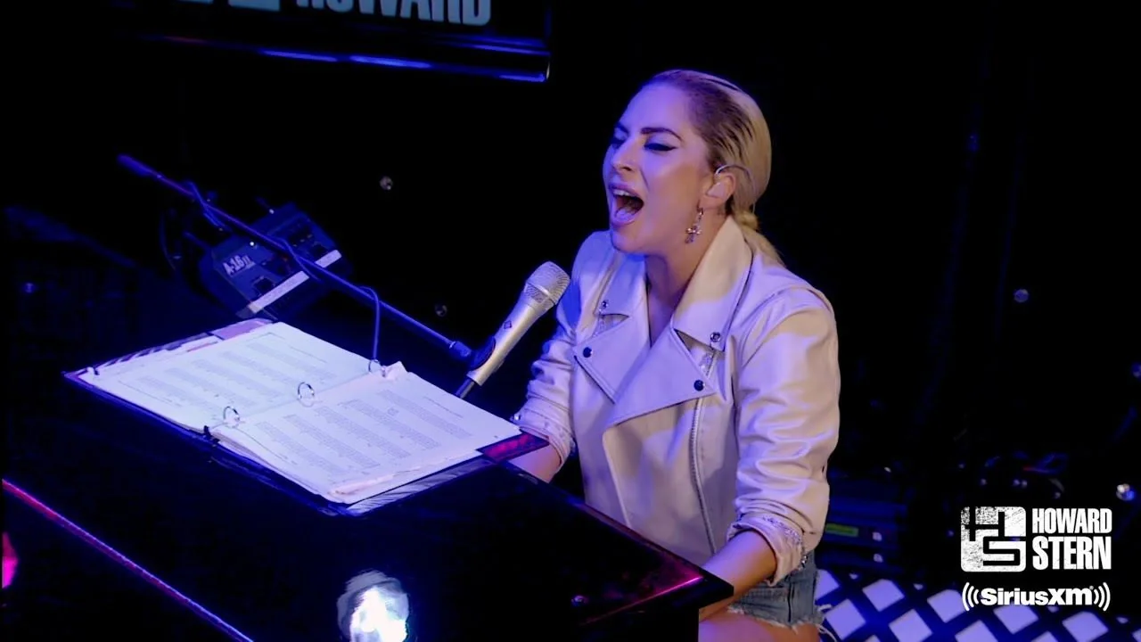Lady Gaga is shown performing in a studio setting, seated at a piano. She is wearing a light-colored jacket, and her hair is styled back in a sleek manner. The atmosphere is intimate, with dim lighting that highlights her expressive singing as she is captured in a moment of passion. A microphone is positioned near her, indicating a live or recorded performance. In front of her on the piano, there is an open binder with sheet music, suggesting she is performing a song. The scene is part of the Howard Stern show on SiriusXM, as indicated by the logos in the lower right corner. The setting is professional, focusing on Lady Gaga's musical talent and emotional connection to the performance. The overall composition emphasizes her as a central figure in the scene.
