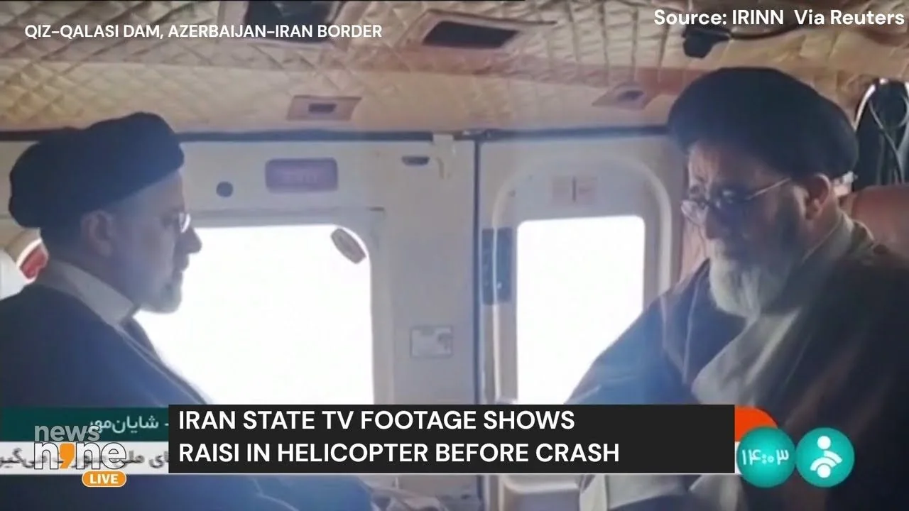 In this video frame, two men are seated inside a helicopter, captured in a somber moment. The man on the left is facing the man on the right, and they both appear engaged in conversation. The setting is inside a helicopter, with quilted padding on the ceiling and visible windows, suggesting mid-flight. Both individuals are wearing traditional attire, including turbans and long coats, indicative of their cultural background. The lighting inside the helicopter is soft, with natural light filtering through the windows.
On-screen text indicates that the footage was aired by IRINN and sourced via Reuters, with the location specified as Qiz-Qalasi Dam on the Azerbaijan-Iran border. The title text informs viewers that this is Iran state TV footage showing Raisi in a helicopter before a crash. The mood seems serious, potentially reflecting the gravity of the situation. The branding and channel information are prominently displayed in the lower third, emphasizing the news aspect of the broadcast.