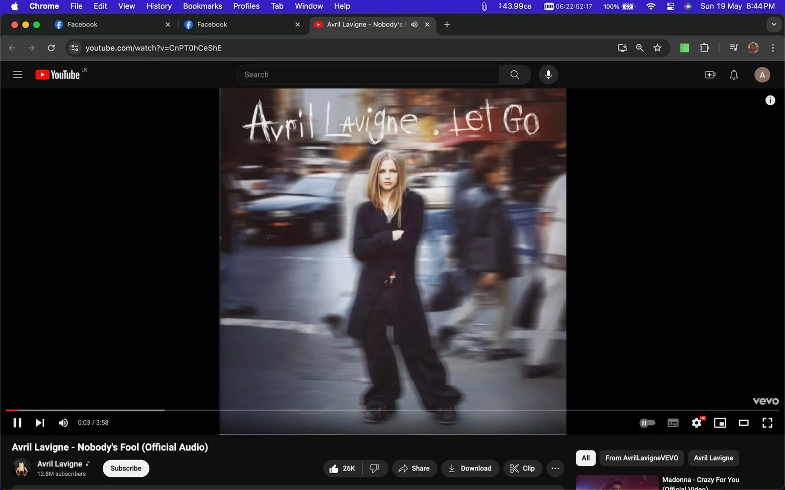 The image is a screenshot from a YouTube video titled "Avril Lavigne - Nobody's Fool (Official Audio)." The channel associated with this content is "Avril Lavigne," and it's published by VEVO. The focal point of the image is the cover of the album "Let Go" by Avril Lavigne. The album cover features a city street scene with Avril Lavigne standing in the center, dressed in dark clothing with her arms crossed, projecting a confident and edgy demeanor. The background is blurred, creating a sense of motion around her as people pass by, highlighting her as the central figure. The colors are muted with a focus on the contrast between her attire and the softer background. The title "Avril Lavigne - Let Go" is prominently displayed in a stylized handwritten font at the top of the cover. The interface includes typical YouTube elements like the play button, video length indicator, and options for liking, sharing, and downloading the video.