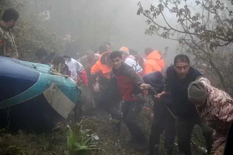 The image depicts a rescue operation at the site of a helicopter crash in a foggy forest setting. Several individuals, including emergency responders in high-visibility orange jackets, are actively engaged in the scene. The responders appear to be assisting or pulling others away from the crash site, showcasing urgency and teamwork. The helicopter's body is partially visible, with its blue and white exterior showing signs of damage. Dense fog envelops the area, adding to the chaotic and tense atmosphere. The ground is covered in dense vegetation, and some responders hold onto branches for support. The lighting is natural but dim, suggesting an overcast day. The expressions on the faces of those present indicate determination and focus on the rescue effort.