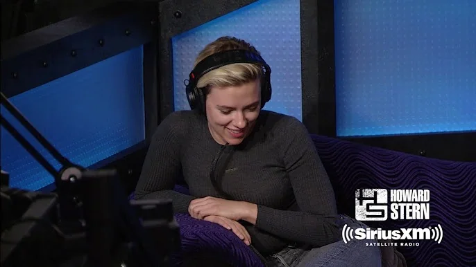 SECTION 2: Rich Prose
The image features a scene from a SiriusXM Satellite Radio broadcast in the Howard Stern studio. A person is seated in a cozy, dark purple chair, wearing large headphones and a fitted dark grey top. She has a pleasant expression, seemingly caught in a moment of laughter or light conversation, adding to the relaxed and informal atmosphere typical of radio interviews. The studio's walls are padded with blue acoustic panels, contributing to the professional and soundproof environment.
Prominent branding for "HOWARD STERN" is visible on the bottom right, alongside the "SiriusXM SATELLITE RADIO" logo, indicating the association with this popular radio show. The background is softly lit, with the blue hues providing a calm and cool ambiance. The overall setting is intimate, focusing on personal interaction, characteristic of Howard Stern's interview style. The well-composed image captures the essence of a broadcast moment, highlighting the engaging nature of live radio.
- Title: "Howard Stern"
- Channel / profile: "Howard Stern"
- Site / app: "SiriusXM"