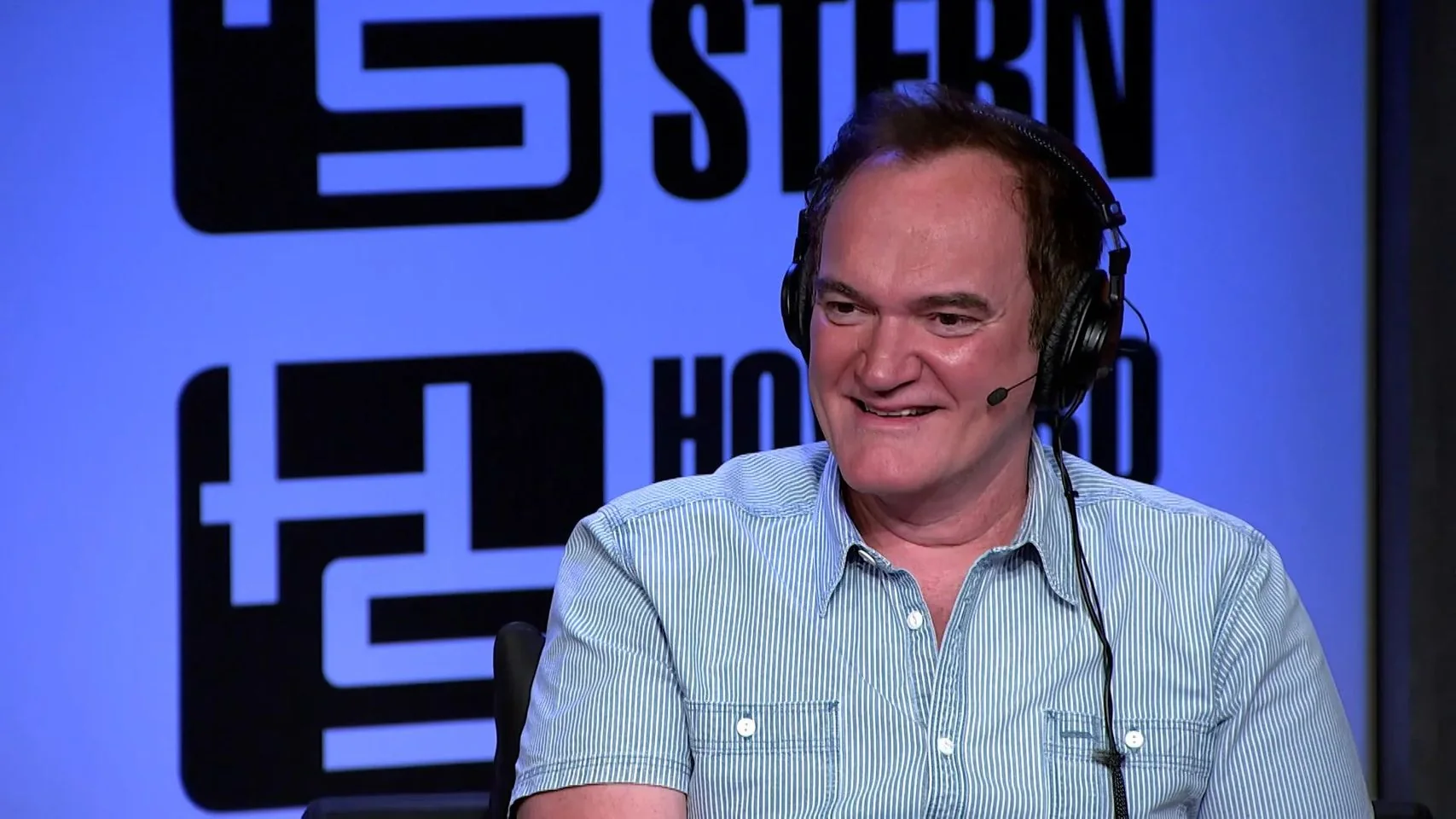Quentin Tarantino is pictured in a studio setting, wearing a light blue, short-sleeved, button-down shirt with white stripes. He appears to be engaged in a conversation, as evidenced by the headset with an attached microphone that he is wearing. The background features the words "STERN" and "HOWARD" prominently displayed, suggesting that this is likely taking place during a recording or broadcast related to Howard Stern. Tarantino has a pleasant expression, indicating he might be responding to a question or engaging in light conversation. The lighting is bright and even, typical of indoor studio environments, which provides clear visibility of his facial expressions and attire. The overall mood of the scene is casual yet professional, fitting for an interview or talk show appearance.