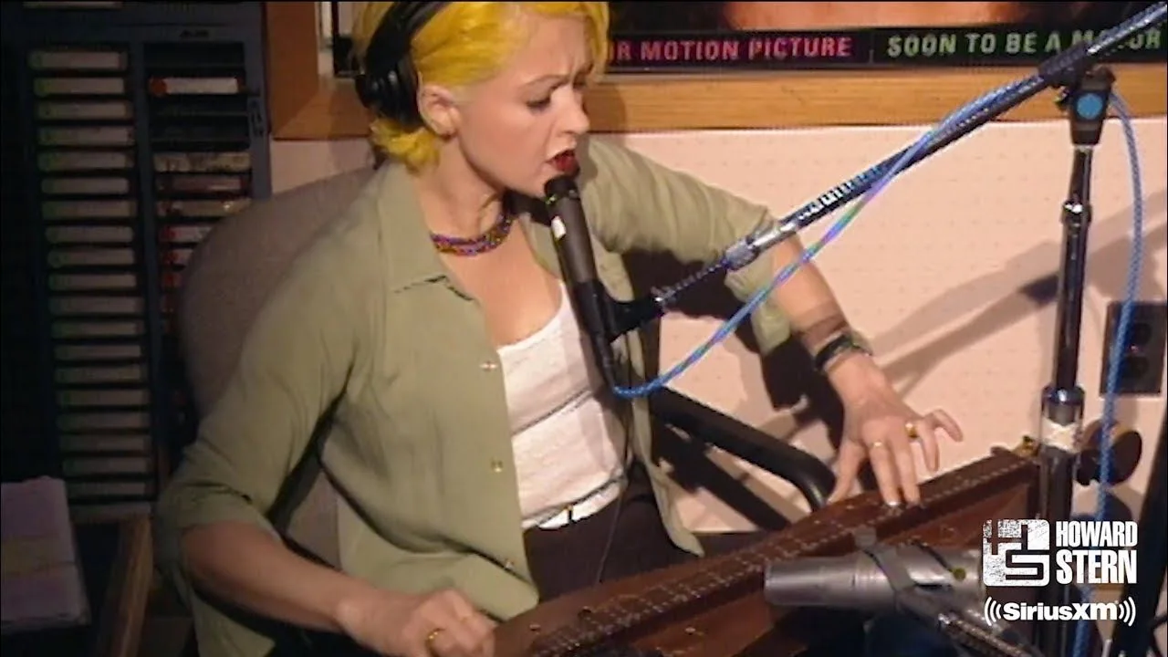 The image shows a radio studio setting, likely part of the Howard Stern show on SiriusXM, as indicated by the on-screen text. A person is seated, engaged in playing a musical instrument that resembles a dulcimer or autoharp. They are focused on the instrument, with their hand positioned over the strings and looking downward, suggesting concentration. The individual wears a light green shirt over a white top, and they have short, bright yellow hair. Their expression appears serious and intent as they perform. The studio environment includes microphones, audio equipment, and soundproofing typical of a radio broadcast setting. The lighting is soft and neutral, focusing attention on the musician. The SiriusXM and Howard Stern logos are prominently visible, reinforcing the context of a live radio performance. The backdrop is filled with audio gear, highlighting the professional setting.