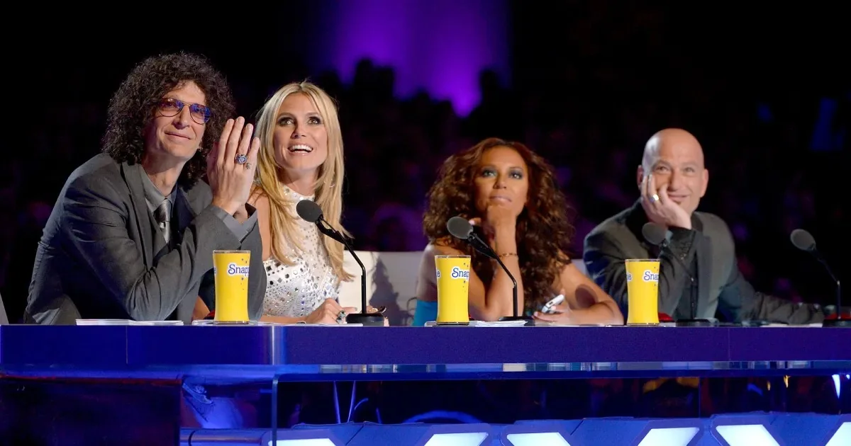 The image shows a panel of judges seated at a table, likely from a talent show such as "America's Got Talent," given the recognizable setting and branding. Howard Stern is featured prominently on the left side. He is wearing a gray suit with a tie and has curly hair. Next to him, a woman with long blonde hair, wearing a white embellished top, is smiling and looking towards the stage. To her right, another woman with voluminous brown hair is resting her chin on her hand, appearing thoughtful. The last person at the table is a bald man, smiling gently with his hand resting on his chin. Each judge has a branded yellow cup in front of them, suggesting product placement for a sponsor. The background is dimly lit with purple lighting, enhancing the moody and dramatic atmosphere typical of a live studio audience setting. The panel is equipped with microphones, indicating they are in the middle of judging or commenting on a performance.
