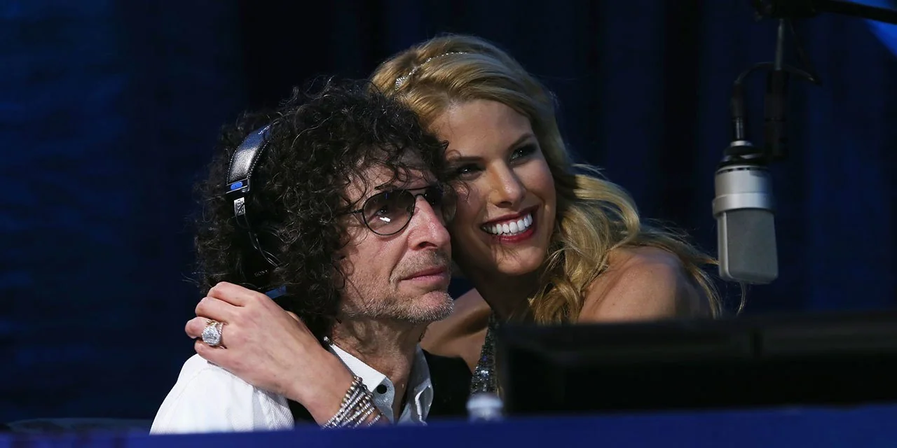 In this image, Howard Stern is seated in what appears to be a radio studio. He is wearing large headphones and glasses, focusing intently on something ahead of him. His clothing consists of a white shirt and a vest, contributing to a professional appearance. Next to him, a woman with long blonde hair is embracing him warmly, smiling brightly towards the camera. Her attire is elegant, suggesting a formal or celebratory occasion. The background is dimly lit, emphasizing the duo in the foreground, while a large microphone is visible, indicating a broadcast setting. The mood is affectionate and lively, as suggested by the woman's cheerful expression and Stern's engaged demeanor. The overall color scheme is dark with highlights, creating a focused and intimate atmosphere typical of a radio studio environment.