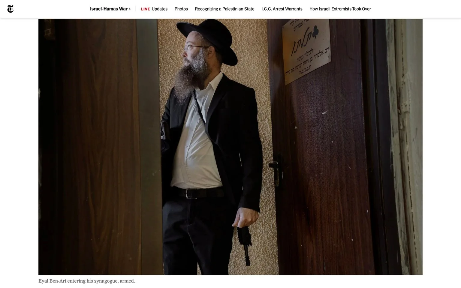 The image shows Eyal Ben-Ari entering a synagogue, as noted in the caption. He is dressed in formal attire, wearing a black suit, a white shirt, and a black hat, which is commonly associated with traditional Orthodox Jewish clothing. Eyal has a long beard and glasses, and he carries a firearm in his right hand. The setting is indoors, with the focus on a door that has Hebrew text on a plaque, indicating the cultural and religious context. The lighting is soft and natural, casting a gentle illumination on Eyal as he stands at the entrance. The background features a textured wall, adding depth to the scene. The atmosphere is serious, reflecting the gravity of carrying a weapon into a place of worship. The image is part of an article from "The New York Times," which is reporting on the Israel-Hamas War, as indicated by the title at the top of the webpage.