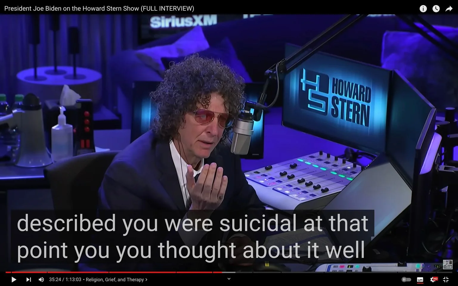 The image is a screenshot from a YouTube video titled "President Joe Biden on the Howard Stern Show (FULL INTERVIEW)." The scene is set in a radio studio, with prominent branding for SiriusXM and the Howard Stern Show. Howard Stern, identified by the provided hint, is speaking into a large, professional microphone. He is wearing a dark suit and red-tinted glasses, and his curly hair is a notable feature. The background is a dimly lit studio environment, with control panels, monitors, and other radio equipment visible. The overall lighting is cool-toned, with shades of blue and purple. The on-screen subtitles read, "described you were suicidal at that point you you thought about it well," which suggests a serious conversation topic. The atmosphere is professional and focused, aligning with the setting of a notable radio broadcast.