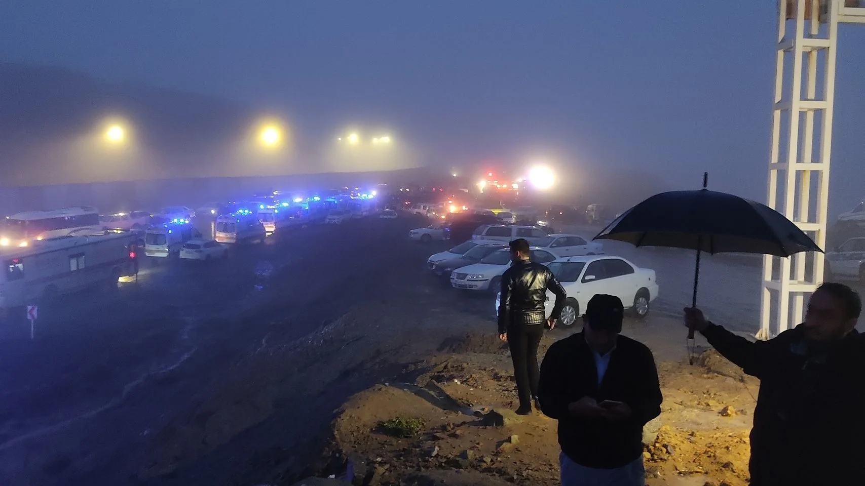 The image depicts a nighttime scene, shrouded in fog, creating a mysterious and tense atmosphere. In the foreground, a person is holding an umbrella, providing some cover from what seems to be wet conditions, suggesting rain or mist. Another individual stands nearby, gazing towards the scene with their hands in their pockets, exuding a sense of waiting or anticipation. The road is lined with a series of vehicles, many of which have their blue emergency lights flashing, indicating a possible emergency situation or traffic incident. The headlights and emergency lights create bright spots in the foggy night.
A substantial number of vehicles are visible, some appearing to be emergency or service vehicles. The road is muddy, hinting at recent rain or poor conditions, contributing to the challenging situation. The background is dominated by bright lights, possibly from more vehicles or streetlights, adding to the visibility difficulties. The overall mood is tense, with the combination of emergency lighting and fog creating an urgent yet obscured scene.