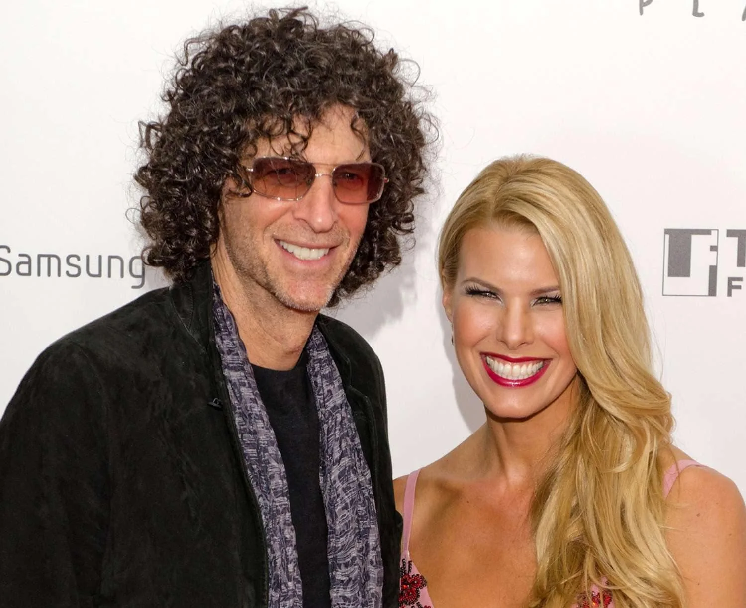 Howard Stern is pictured at a red carpet event alongside a woman with long blonde hair. Stern is wearing a black jacket over a dark shirt, accessorized with a patterned scarf and sunglasses. He has a pleasant expression, with his signature curly hair adding to the relaxed vibe. The woman beside him is wearing a pink dress with intricate detailing and has a bright, wide smile that exudes confidence and happiness. The background features a white backdrop with logos including "Samsung" and "FT," indicating a formal event or premiere. The lighting is even and clear, typical of red carpet settings, highlighting the subjects against the branded backdrop. The overall atmosphere is lively and glamorous, capturing a moment of celebrity engagement.