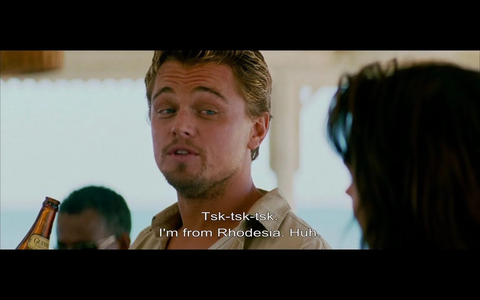 The image is a screenshot from the film "Blood Diamond," featuring a character portrayed by a well-known actor in an outdoor setting. The scene depicts a conversation between two characters, with the actor facing the camera and another character partially visible from behind. The setting suggests a casual atmosphere, possibly on a veranda or outdoor bar, given the presence of natural light and a relaxed posture. The actor is wearing a light-colored shirt, slightly unbuttoned, exuding a laid-back yet confident demeanor. He is holding a bottle that appears to be a beer, with a visible label, indicating a casual social setting.
His expression is slightly smug or amused, complementing the subtitle dialogue: "Tsk-tsk-tsk. I'm from Rhodesia. Huh." This line suggests a moment of self-identification or perhaps a retort within the conversation. The lighting is bright, indicative of daytime, enhancing the scene's vibrant and natural look. The composition is focused on the actor, with other background elements slightly out of focus, drawing attention to the dialogue and character interaction pivotal to the scene. This setting and interaction are characteristic of "Blood Diamond," where themes of identity and conflict are central.