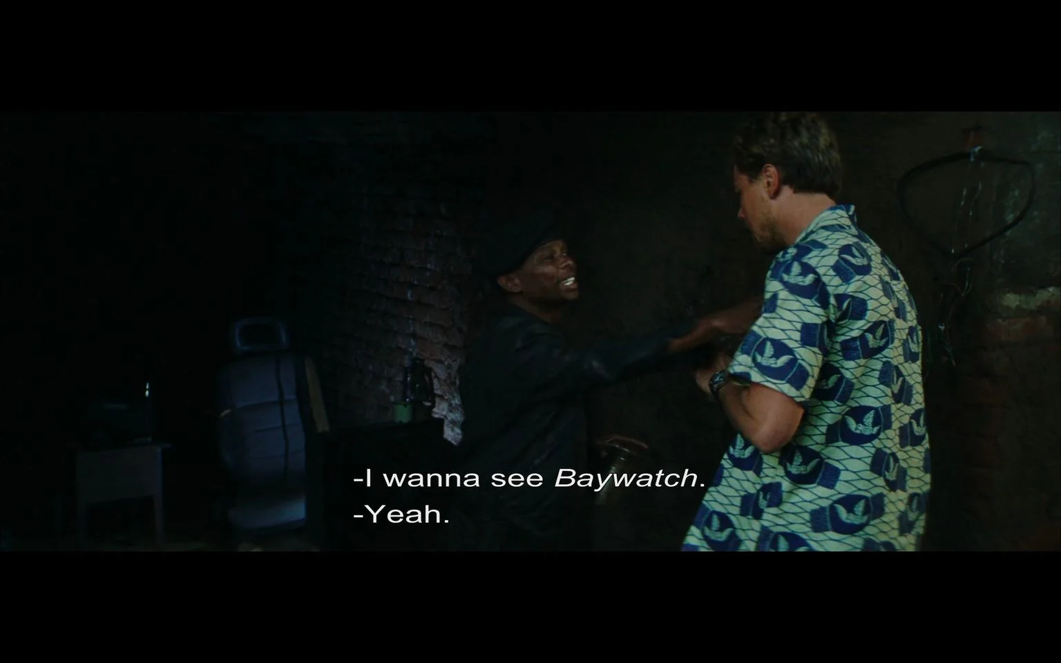 The image is a scene from the film "Blood Diamond," featuring a conversation between two characters in a dimly lit, indoor setting. The man on the left is wearing a dark jacket and hat, animatedly gesturing with his hand, while the man on the right, partially visible, is wearing a patterned shirt with shades of blue and white. They appear to be in a basement or similarly rustic environment, indicated by the exposed brick wall and sparse furnishings, including a chair and some equipment in the background. The lighting is low, creating a tense or secretive atmosphere. The mood is casual yet intense, as reflected in the dialogue where one character expresses a desire to watch "Baywatch," to which the other responds affirmatively. The composition focuses on the interaction between the two characters, highlighting their expressions and body language.
- Captions / subtitles: "- I wanna see Baywatch.","- Yeah."
