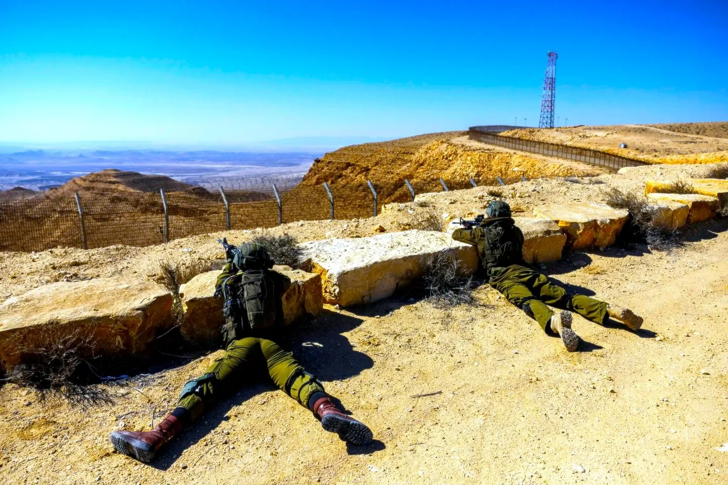 Two soldiers are positioned in a desert setting, likely on a border patrol or training exercise. They are lying prone on the ground, partially sheltered behind large rocks. Each soldier is aiming a rifle towards the expansive desert landscape, which stretches out into the horizon. The terrain is rugged with hills and a clear blue sky overhead, suggesting a dry and arid climate. A fence runs parallel to their position, indicating a boundary or perimeter. In the distance, a tall communication tower is visible, adding to the sense of a monitored area. The soldiers are wearing standard military gear, including helmets and backpacks, and their uniforms are a muted green, suitable for blending into the environment. The overall scene is calm yet tense, reflecting readiness and vigilance.