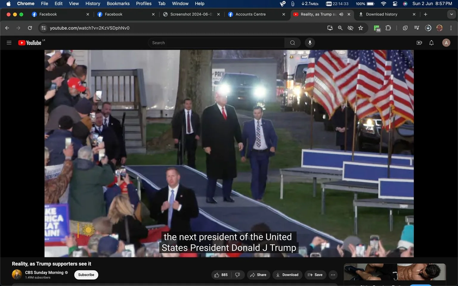 This image is a screenshot from a YouTube video titled "Reality, as Trump supporters see it" on the CBS Sunday Morning channel. It captures an outdoor scene at what appears to be a political rally. In the foreground, a crowd of supporters is visible, many holding up phones to capture the moment. The background shows a prominent figure, who, based on the context, is Donald J Trump, walking down a ramp flanked by men in suits, possibly security personnel. The setting includes several American flags displayed prominently to the right, contributing to the rally atmosphere. The lighting suggests early evening or nighttime, with visible headlights from a vehicle in the background. The mood is energetic, with people in the crowd actively engaged, holding signs and phones. The video’s caption at the bottom reads, "the next president of the United States President Donald J Trump," highlighting the focus on the political event and its significance to the supporters gathered there.