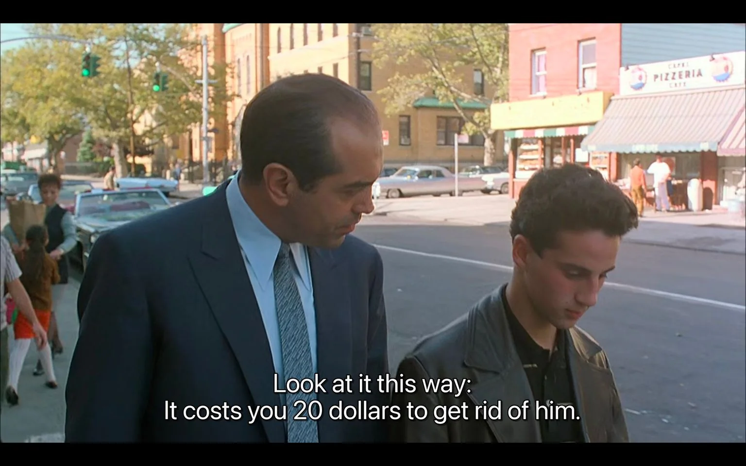 The image is a scene from the film "A Bronx Tale." It depicts two characters walking along a street during the day. The man on the left is dressed in a formal dark suit and tie, looking down at the young man beside him. The younger character is wearing a leather jacket and looking down, seemingly contemplative or concerned. The setting is an urban neighborhood with a visible pizzeria in the background, along with parked cars and pedestrians, suggesting a lively street scene. The lighting is bright and natural, typical of daylight. The mood conveyed through their expressions and body language suggests a serious or instructional conversation. The subtitles at the bottom of the image read, "Look at it this way: It costs you 20 dollars to get rid of him," indicating the nature of the dialogue. The style is cinematic, fitting for a dramatic narrative sequence.